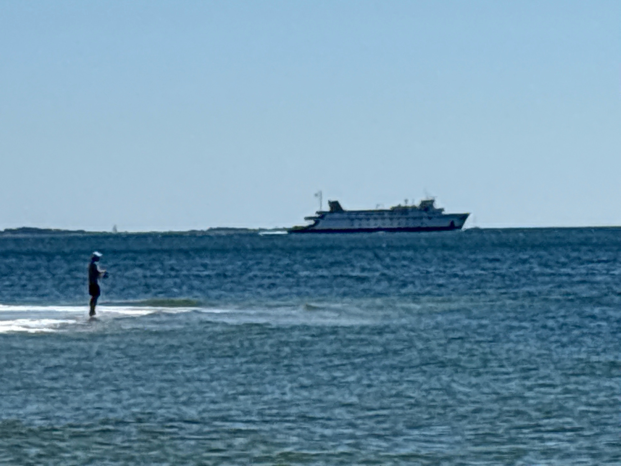 A wide-angle, full shot captures a serene maritime scene on a bright, clear day. In the foreground, a person stands in the water, appearing to be fishing. The individual is silhouetted against the bright water, wearing a hat and shorts, with their arm extended as if casting a line. The water itself is a deep blue, with gentle waves and a light foam trailing behind the person, suggesting shallow water or a sandbar.
In the mid-ground, a large ferryboat sails horizontally across the horizon. The ferry is dark in color and appears to be a passenger or car ferry. Its distinct profile with multiple decks and a superstructure is visible.
The background is dominated by a clear, light blue sky. A faint, dark line on the horizon likely represents land or a distant coastline. There is also a small, white triangular shape, possibly a distant sailboat, visible on the far left horizon. The overall impression is one of a peaceful day at sea, with human activity contrasted against the vastness of the water and sky. The image is of the Waterford, United States of America.