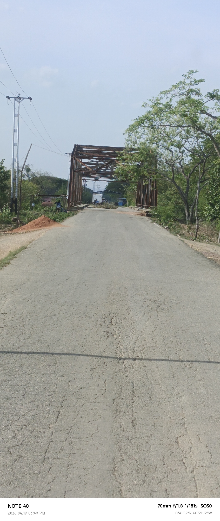 The image shows a weathered asphalt road leading towards a metal bridge structure. The bridge appears rusty and possibly old, with trees and vegetation visible on both sides of the road and around the bridge. A utility pole stands to the left of the road near the entrance to the bridge. In the distance, beyond the bridge, there are some structures and vehicles. The road itself is cracked and shows signs of wear. The image was taken at 8°41'59"N 68°21'12"W.