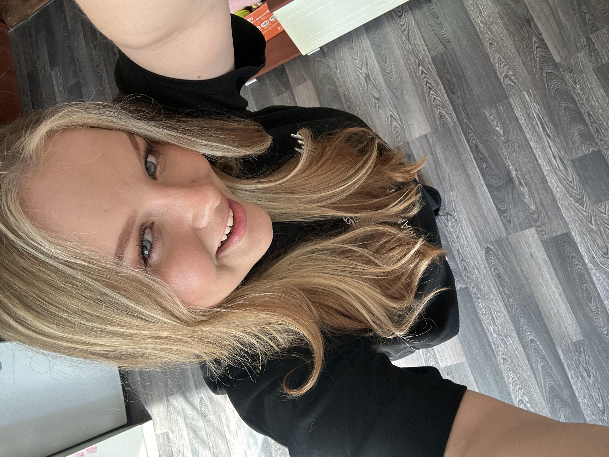 A blonde-haired woman in a black hooded sweatshirt is pictured from a high-angle perspective. The floor is grey laminate with wood grain. A few objects are visible out of focus in the background. The location of the image is not known.