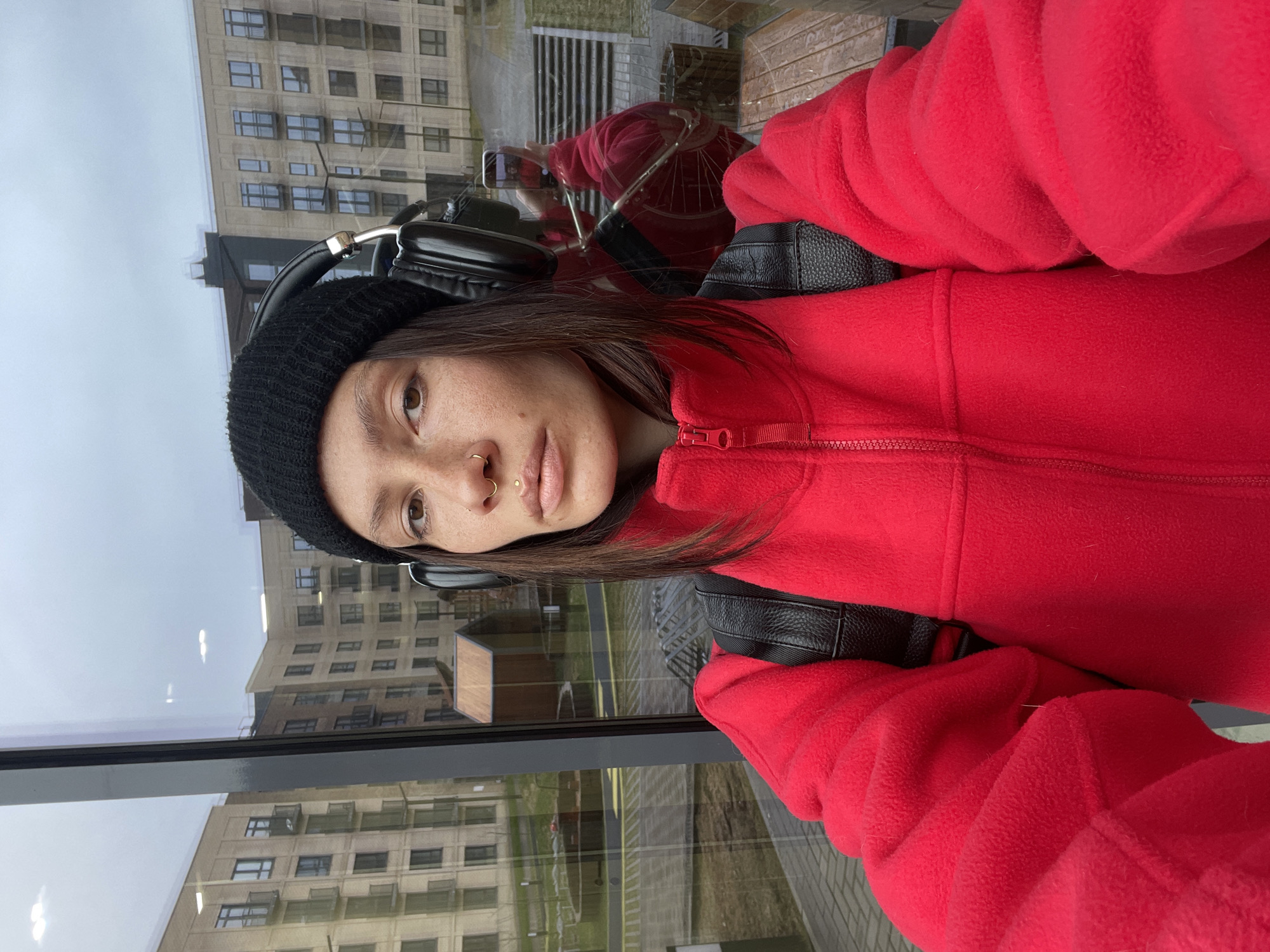 A young woman is taking a selfie indoors, looking out through a large window, likely in Pervomaysky, Russia. She is wearing a bright red fleece jacket, a black knitted beanie, and black over-ear headphones. Her dark hair frames her face, and she has several facial piercings, including a gold septum ring, a gold nostril ring, and a gold philtrum piercing. Her expression is neutral as she looks directly at the camera. Visible black backpack straps are worn over her red jacket.
Through the window, an urban or suburban landscape is visible under an overcast, light gray sky. Tall, multi-story buildings with numerous windows, constructed from light-colored materials, can be seen. The ground outside features patches of green grass and some bare earth or pavement. Reflections on the glass surface show the woman holding a smartphone, confirming the selfie action. The diffused light suggests it is daytime, and the warm clothing (fleece jacket, beanie) indicates cool weather. No discernible text is visible in the image.
