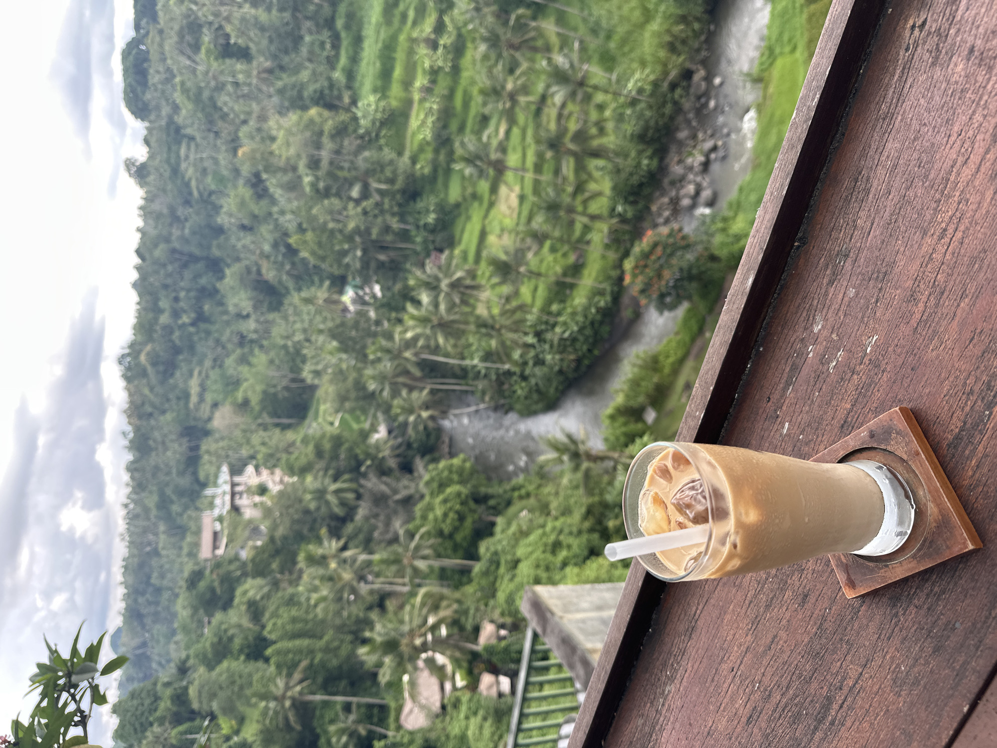 On a wooden surface, a glass of iced coffee rests on a square coaster. In the background, a lush, green landscape unfolds, featuring terraced rice paddies, dense vegetation, and a winding river. Palm trees are interspersed throughout the landscape. The location is Ubud District, Indonesia.