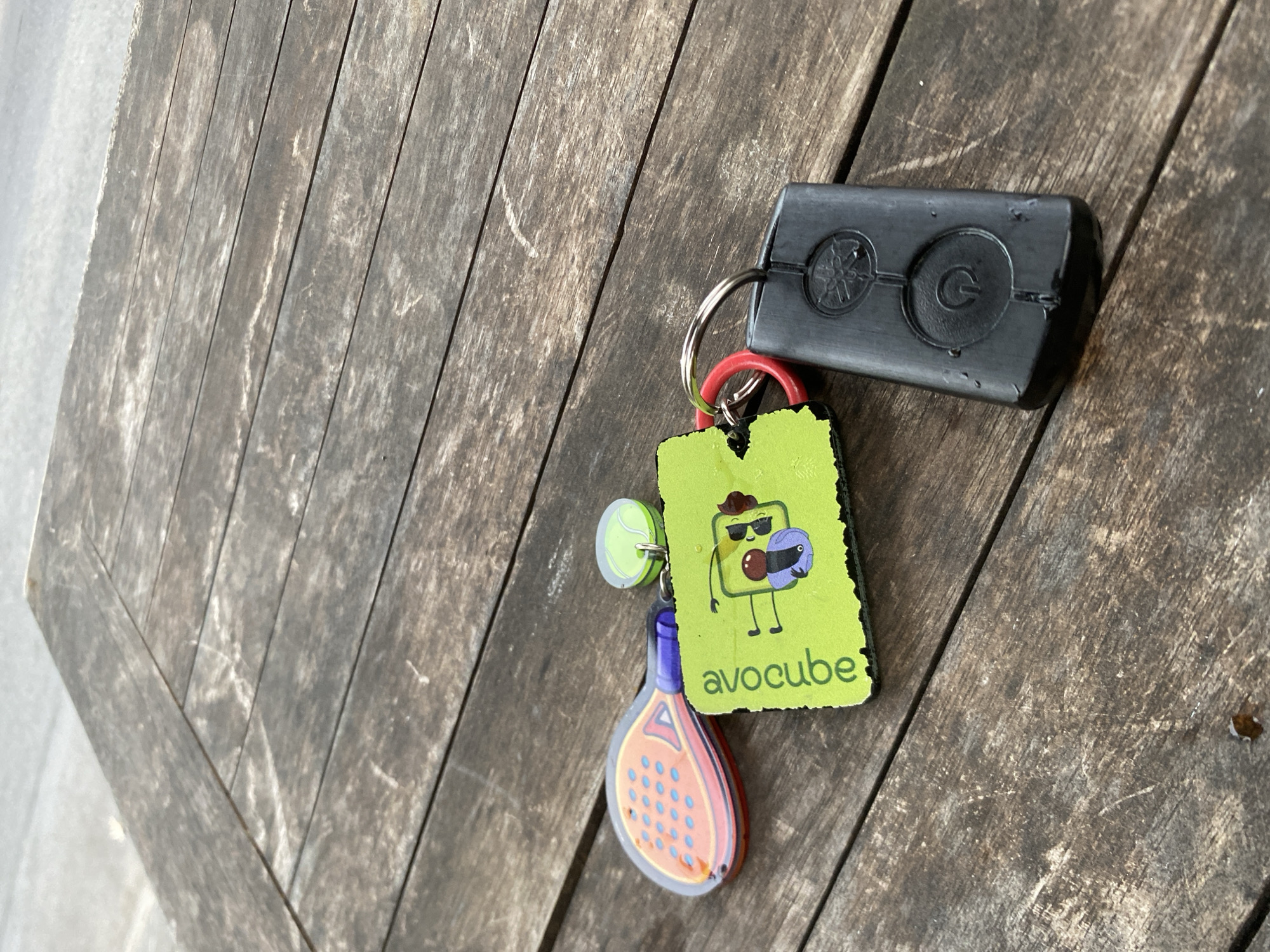 A set of keys and two small novelty charms are resting on a weathered, slatted wooden surface, likely a table. One charm is shaped like a miniature orange and pink tennis racket with blue dots, attached to a purple plastic handle. The second charm is a bright green square with a cartoon character that resembles a cube with arms and legs, sunglasses, and a purple bow tie. The word "avocube" is written vertically in white text on this charm. Attached to the charms is a black, rectangular electronic device, possibly a key fob for a car or security system, with a circular power symbol embossed on its surface. A silver key ring connects all these items, with a small red ring also visible. The lighting suggests an outdoor setting.