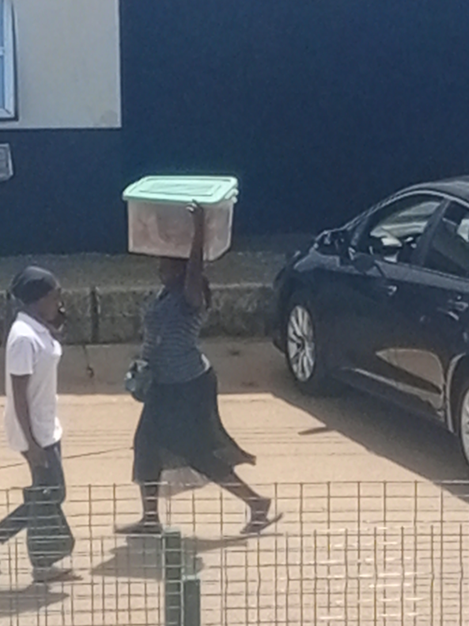 In Bukuru, Nigeria, two individuals are present on a sunlit street. The primary subject is a woman walking from right to left, balancing a light green plastic container on her head. She wears a striped shirt and a dark, flowing skirt, with dark shoes. A dark bag hangs from her shoulder. To her left, a person wearing a white short-sleeved shirt and dark pants stands facing her, their head covered with a dark headscarf or hat. A dark-colored car is parked or moving on the right side of the frame, partially obscuring the background. The scene appears to be near a building with a light-colored wall and a dark blue facade. A wire fence runs across the foreground, casting shadows from the individuals onto the ground. The ground is paved and appears dusty.