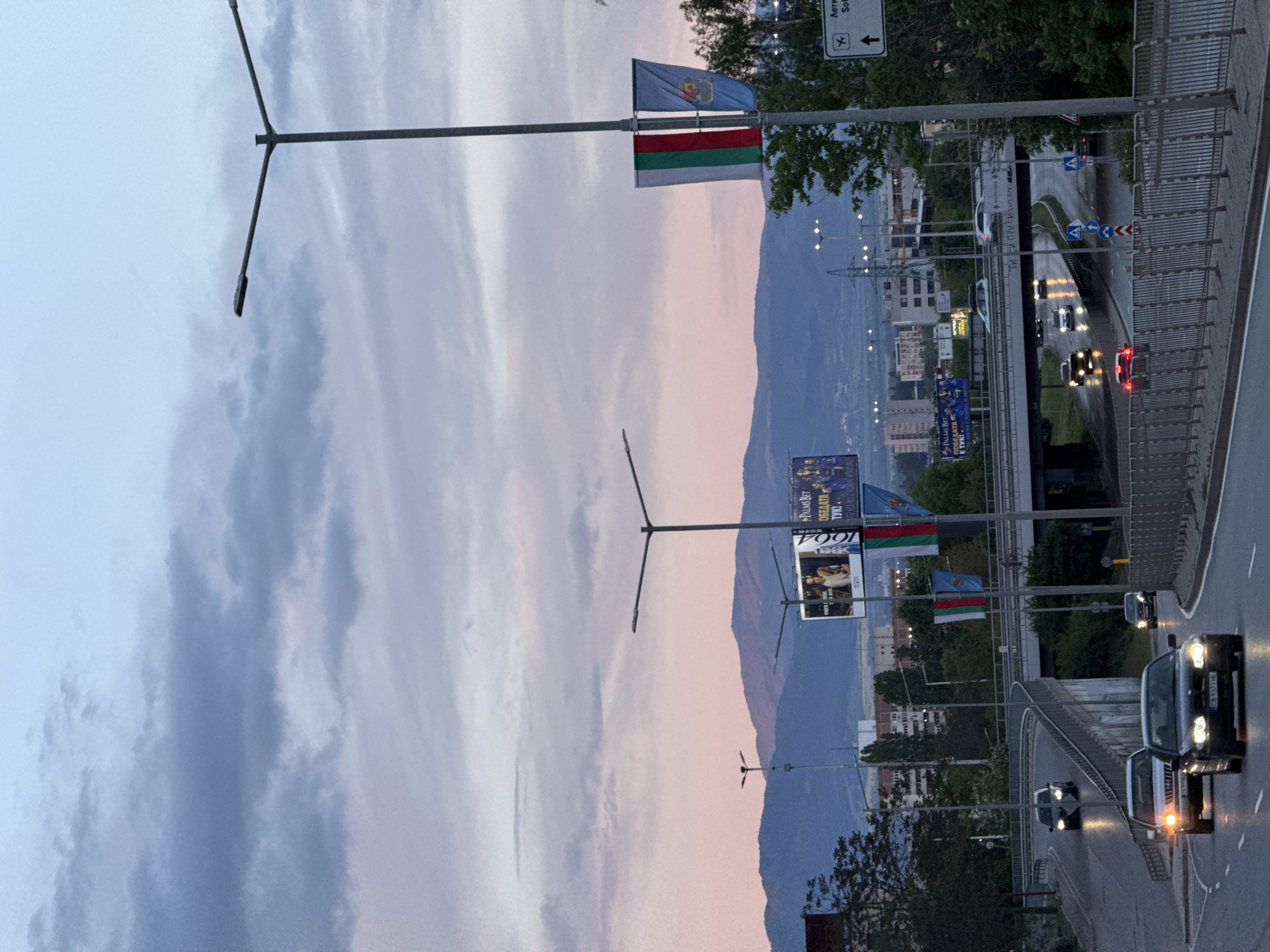 This image captures an urban landscape in Sofia, Bulgaria, during dusk or early evening. The sky is a prominent feature, displaying soft gradients of blue, pink, and purple, with visible cloud formations stretching across.
In the foreground, a multi-lane road or highway is visible, populated by several cars, some with their headlights and taillights illuminated, indicating the low light conditions. Metal barriers line the elevated road structure. Modern streetlights with distinctive three-pronged designs illuminate the area.
Multiple flags are displayed on poles along the road. The national flag of Bulgaria (white, green, and red horizontal stripes) is clearly visible, often accompanied by a blue flag featuring an emblem, likely representing the city of Sofia. Large advertising billboards stand alongside the road; one prominently displays "PALMS BET" and the Cyrillic text "ПОБЕДАТА Е ТУК!" (The victory is here!) along with the number "1604." A white street sign, partially visible, indicates "Лети Со," likely referring to "Летище София" (Sofia Airport), providing a clear location cue.
In the midground, various buildings contribute to the cityscape. The background features a range of mountains, their forms softened by the twilight, contrasting with the urban elements. The overall scene depicts a busy city thoroughfare at a transitional time of day, with both man-made structures and natural topography visible.