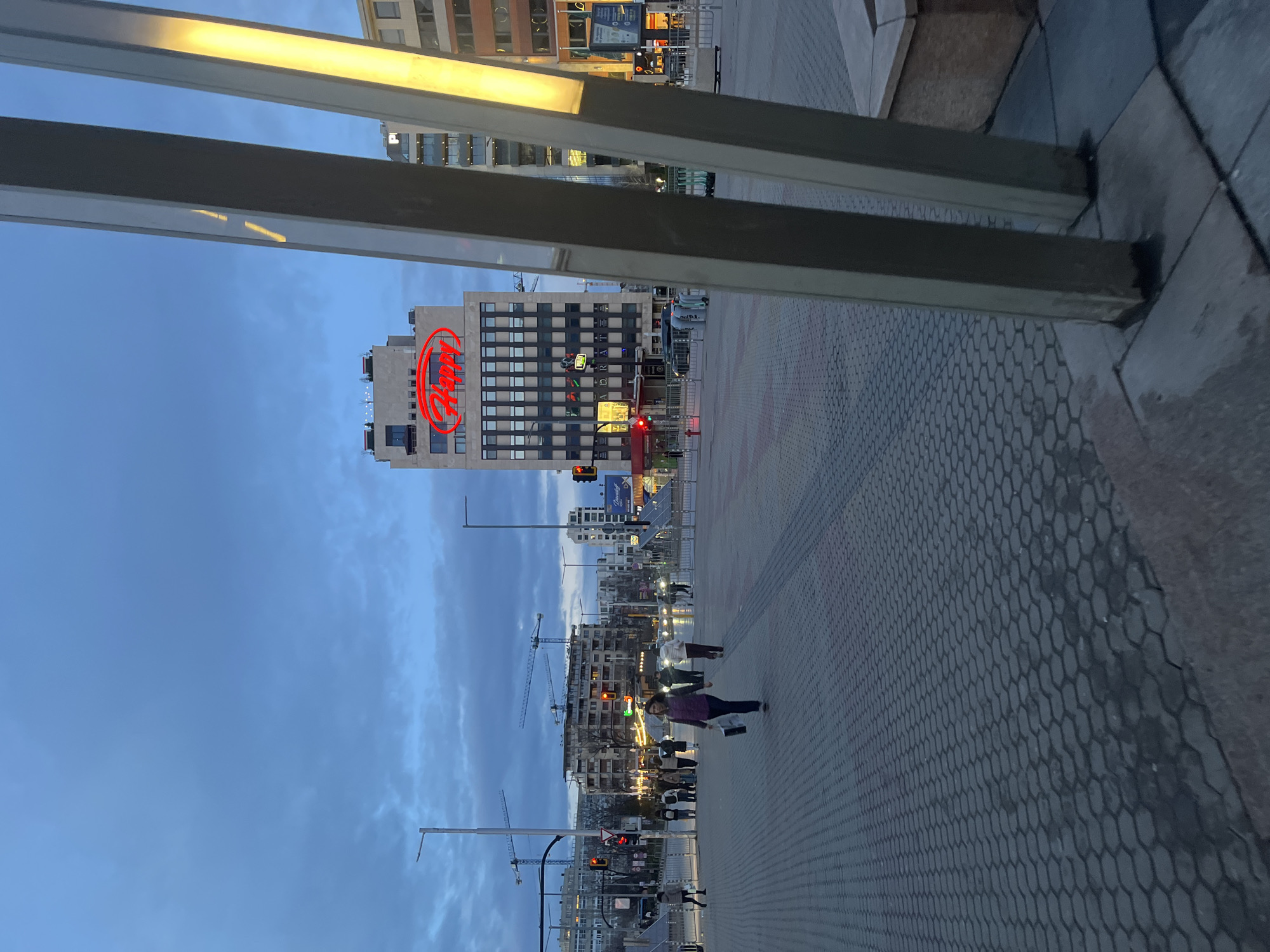 The image depicts a cityscape, likely in Sofia, Bulgaria, judging by the distinctive red "Happy" restaurant sign atop a high-rise building. The scene is viewed from a low vantage point, framed by a metallic structure. The sky is a blend of blue and twilight hues. Pedestrians are walking on a patterned brick walkway that makes up a significant portion of the foreground. Construction cranes are visible further in the background, and multiple buildings are visible, many with illuminated windows. Streetlights and traffic signals add to the urban environment.