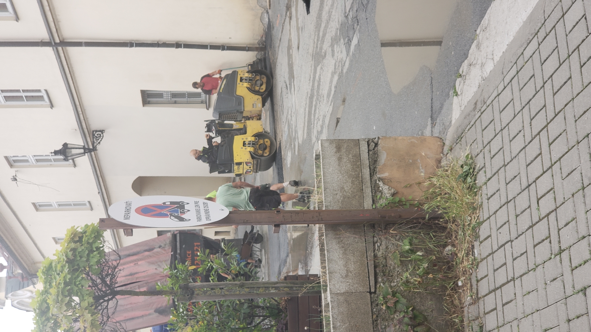 The image captures a street scene in Prešov, Slovakia, likely during road repair work. In the foreground, a paved sidewalk is visible with a concrete planter box. A tall metal pole with a sign is mounted in the planter box. The sign is circular with a red border and a diagonal red line across a red circle, indicating a prohibition. Below the prohibition symbol, text reads "NEPARKOVAŤ!" (Do not park!) and "PARKOVANIE LEN PRE OPRAVNENÉ OSOBY" (Parking only for authorized persons).
Behind the sign, a person wearing a green t-shirt and black shorts is standing, facing away from the camera. To their right, two road workers are operating a yellow asphalt roller. One worker, wearing a red shirt, operates the roller from a seated position, while another worker, in a dark jacket, stands and appears to be guiding or assisting. The roller is compacting asphalt on the street.
In the background, a large truck with a red tarp is parked, and part of a building with cream-colored walls and windows is visible. A street lamp is mounted on the building wall. The street surface appears wet in places, suggesting recent rain or the use of water during roadwork. Vegetation is growing in the planter box and along the side of the street. The overall atmosphere suggests an active construction or maintenance site within an urban environment.