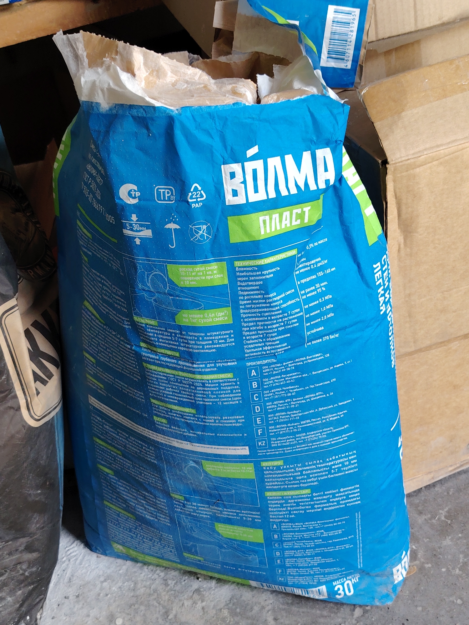 The image shows a large, blue bag of building material, possibly plaster or putty, with Russian text and Cyrillic lettering. The bag is branded "ВОЛМА ПЛАСТ" and has various technical specifications and instructions printed on it. There are several logos and symbols, including a TP certification mark and a recycling symbol. The bag appears to be partially open at the top, revealing some of the powdery contents. It is leaning against a cardboard box in what looks like an indoor storage area, possibly a construction site or a warehouse. The lighting suggests it is daytime, but the environment appears somewhat dimly lit and dusty. There is no visible presence of people, animals, or any specific landmarks that would indicate the city of Oral, Kazakhstan.