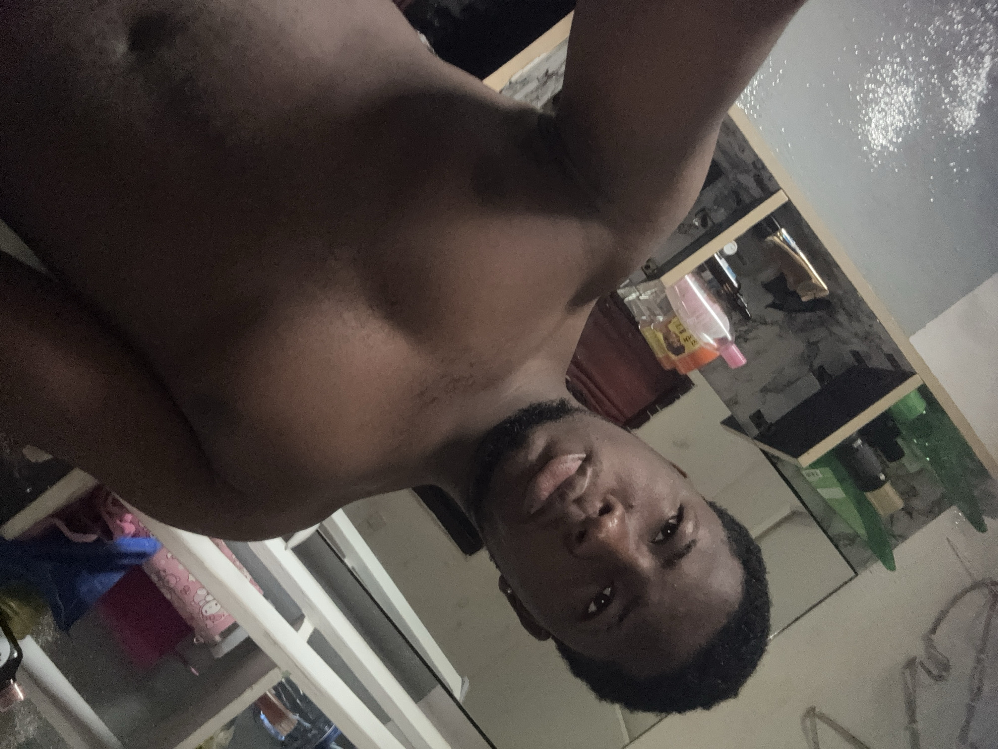 The image is a selfie taken by a young man indoors in Ipetumodu, Nigeria. The man, who has dark skin, short dark hair, and a beard, is shirtless and is looking directly at the camera with a casual, slightly smiling expression, with his tongue briefly visible. The image appears to be rotated 90 degrees counter-clockwise.
The background shows elements of a personal space, likely a bedroom. To the upper left (when viewed in correct orientation), a white structure, possibly a bed frame or shelving, is visible. Beneath it, a pink item adorned with cartoon-like characters (resembling Hello Kitty) and a blue fabric item are seen. To the upper right, a light-colored surface is covered with scattered, reflective white specks, possibly glitter or dust. Below this surface, a shelf or counter holds several bottles, including a prominent pink one and another containing an orange/yellow liquid, suggesting toiletries or household supplies. Other dark objects are on shelves further in the background. The lighting in the room is dim, indicating an indoor setting possibly illuminated by artificial light. No legible text is visible on any objects.