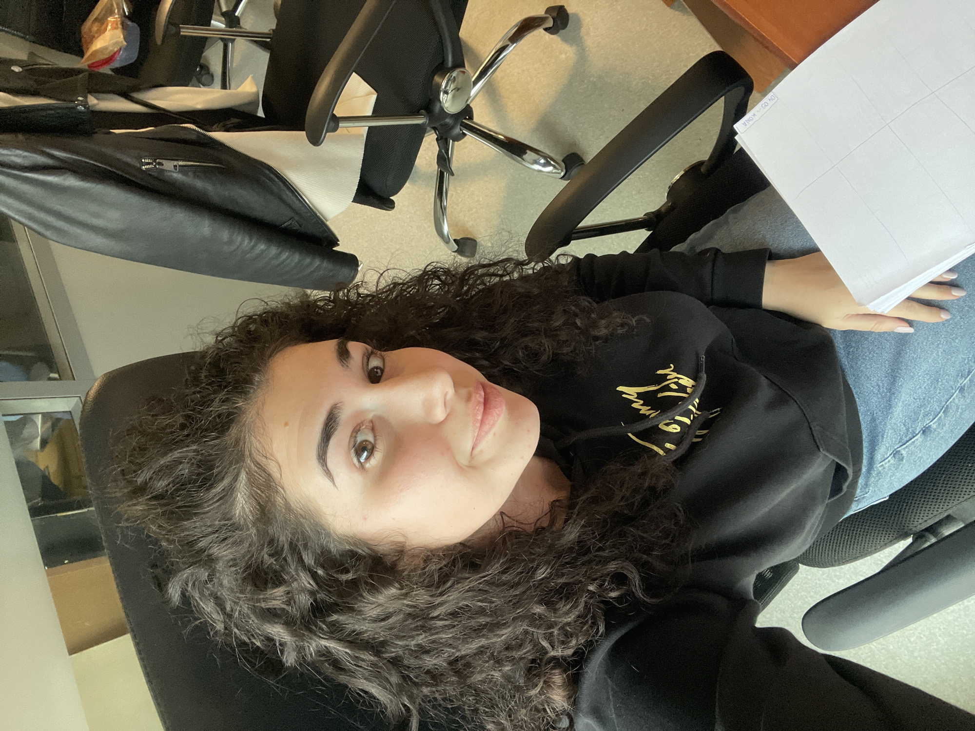 A young woman is depicted in a selfie, looking directly at the camera with a calm, slightly smiling expression. She has dark, voluminous curly hair and brown eyes. She is wearing a black hooded sweatshirt with stylized gold lettering on the front, and blue jeans. She is seated in a black office chair, and in her left hand, she holds a stack of white papers featuring grid lines. Her nails are painted a light, neutral color.
The setting appears to be an indoor office or study space. Behind her, another black office chair with a chrome base is visible, alongside a black leather jacket draped over a chair. A wooden desk surface is also partially seen in the background. The floor is a light, neutral color.
Notable visible text includes "04.03. KONA" printed on the top sheet of the papers she is holding. The gold lettering on her hoodie is partially obscured and not fully legible.