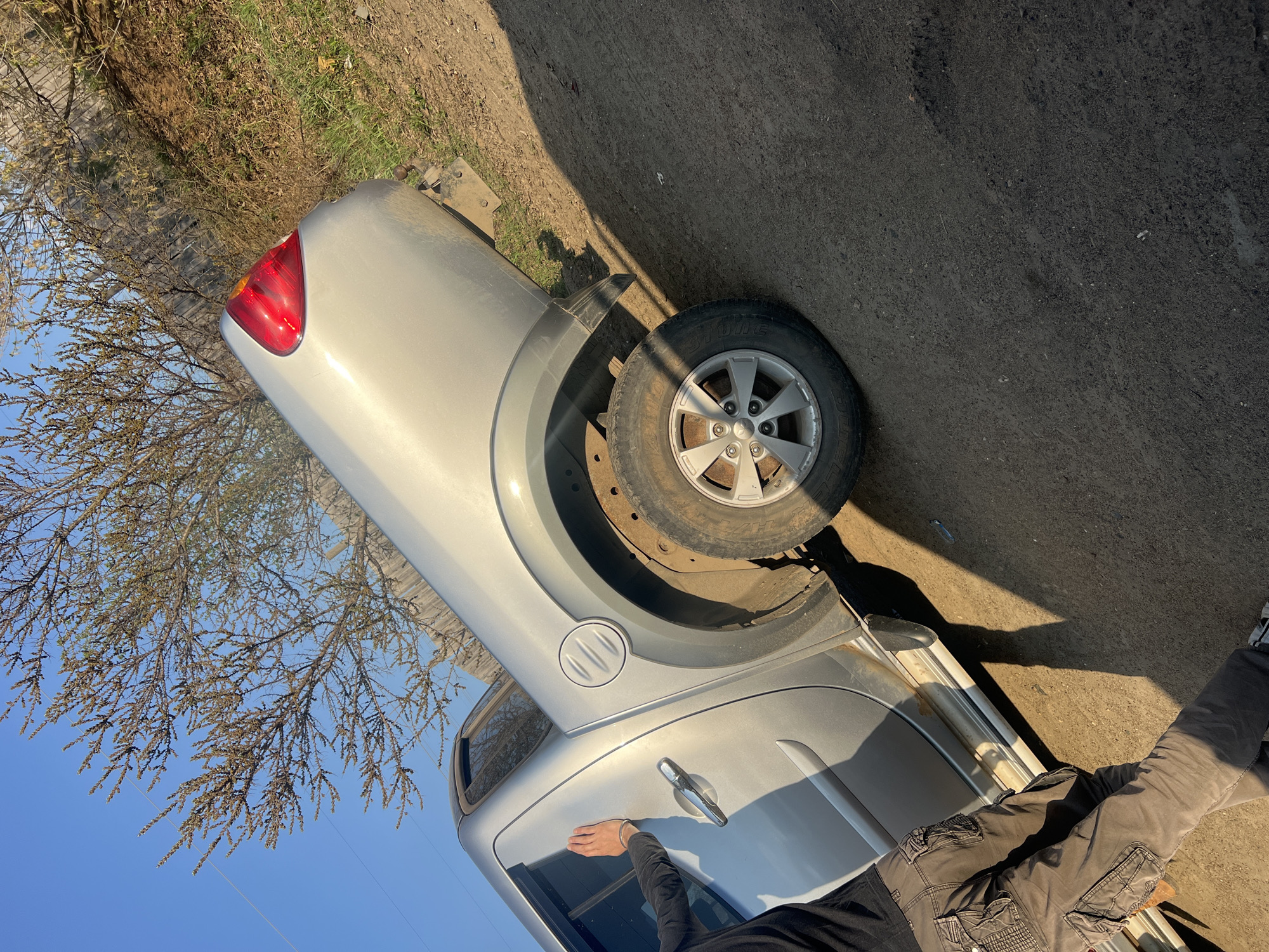A silver vehicle is partially visible in an outdoor environment. The rear section, including a wheel and taillight, is prominently displayed. A person's arm and legs are also visible near the car. The ground appears to be a mix of dirt and asphalt. Branches of a tree are visible. Without additional information, it's impossible to confirm the exact location in Chaikovsky, Russia.