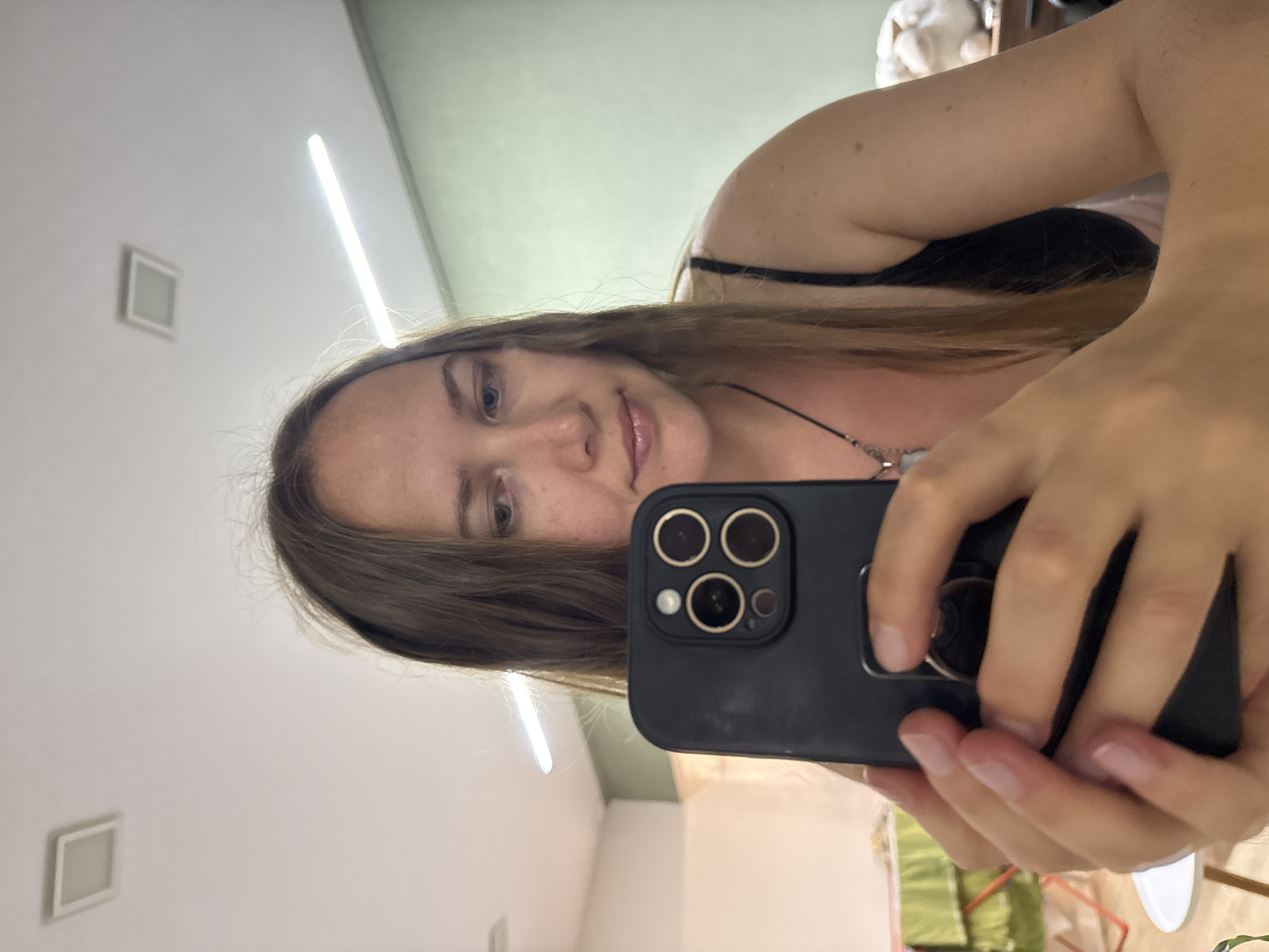 A young Caucasian woman is taking a selfie in what appears to be an indoor setting. She has long, straight, light brown hair and is looking down at her phone, which she is holding in her hands. She is wearing a black tank top and a necklace with a pendant. The background shows a wall, a ceiling with lights, and some furniture, suggesting it might be a home or a studio. There is no information in the image that would allow for identification of the specific location within Izhevsk, Russia.