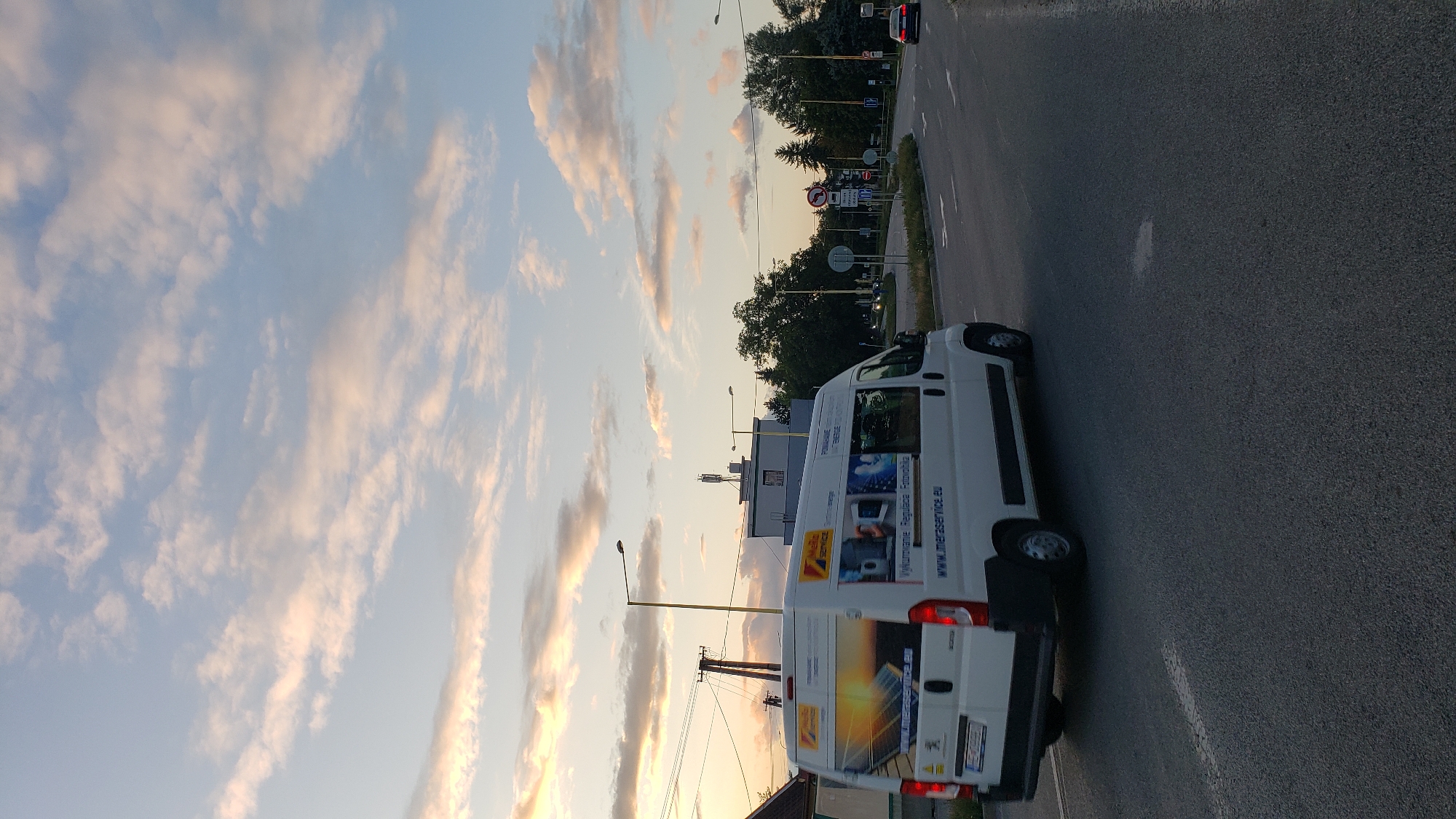 The image captures a street scene at dusk in Prešov, Slovakia. A white van, branded with "Mera Service" and featuring imagery of solar panels and technology, is driving on the right side of a paved road. The van has a license plate that is partially visible. In the background, trees, streetlights, and buildings, including one with a cellular tower on its roof, are present. The sky is filled with scattered clouds, tinged with the warm colors of sunset. Traffic signs are visible along the roadside. A dark-colored car is also seen in the distance on the same road. The overall environment suggests an urban or suburban setting on the outskirts of the city.