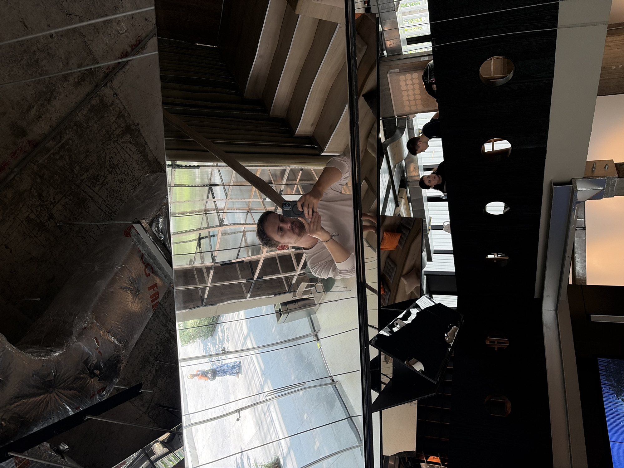 The image captures a multi-faceted reflection, likely taken in Bangkok, Thailand, given architectural styles and general environment depicted. The primary subject is a person holding a smartphone, apparently taking a picture or video of themselves in the reflective surface. The reflections reveal several distinct environments.
On the left, there is a view into what appears to be an unfinished or industrial space with exposed ductwork and rough construction. Adjacent to this, a modern architectural space is reflected, showing metal-framed windows, an outdoor area with greenery, and people.
Further along the reflection, a wooden staircase is visible, as well as other occupants. Another reflection shows elements of a building's interior, specifically a decorative black wall with circular cutouts. This suggests a modern design.
The overall impression is a complex visual tableau where reflections of different spaces and activities are juxtaposed, offering fragmented glimpses of Bangkok's diverse urban settings.