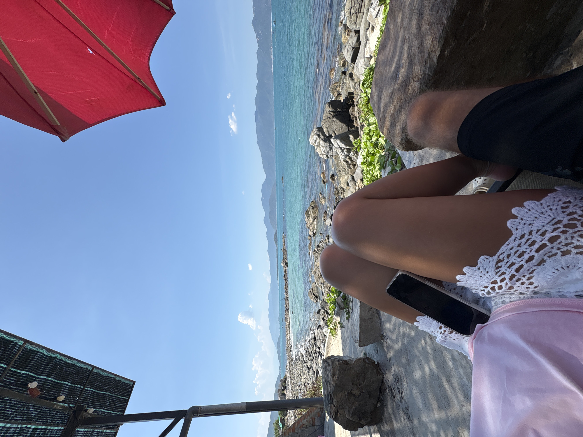 The image shows a beach scene, presumably in Vĩnh Lương, Vietnam, as indicated by the prompt. A person is sitting under a red umbrella, visible in the upper left corner, on what appears to be a coastal area with rocks and vegetation. Another person wearing black shorts is also seated nearby. The sea is visible with a mountainous coastline in the distance under a clear blue sky. A smartphone is positioned between the legs of the first person, who is wearing a white crocheted garment over a pink garment. Part of a shade structure is visible in the lower left corner.