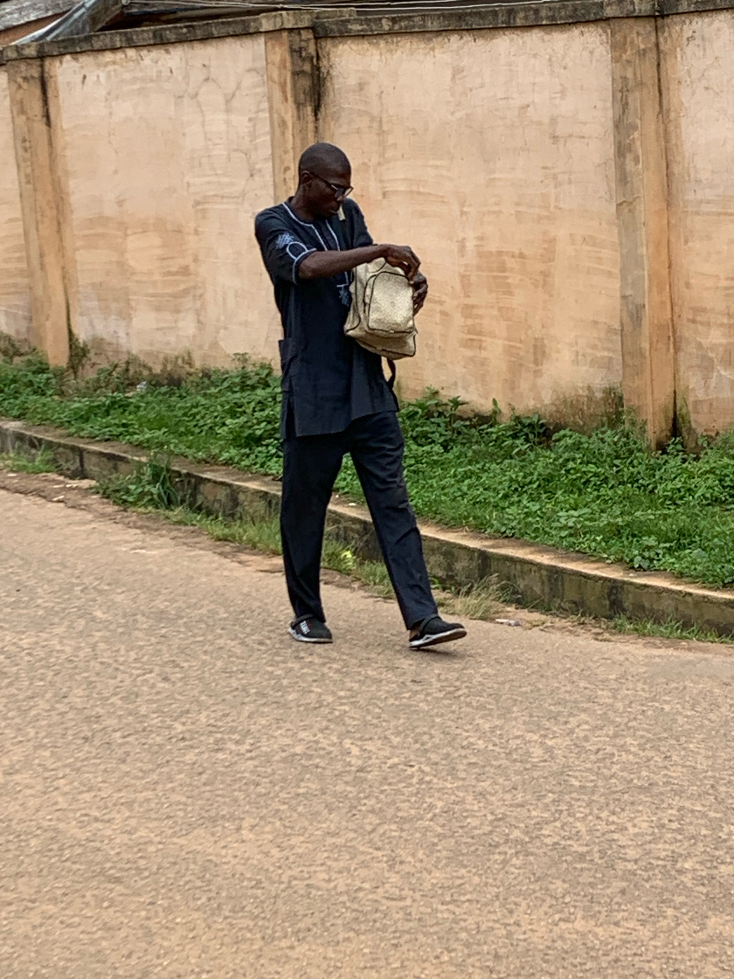 A man walks along a paved road in Jos, Nigeria, towards a concrete wall. He is wearing a dark blue traditional outfit and black sneakers. He is holding a light-colored bag. Lush green vegetation lines the edge of the road.