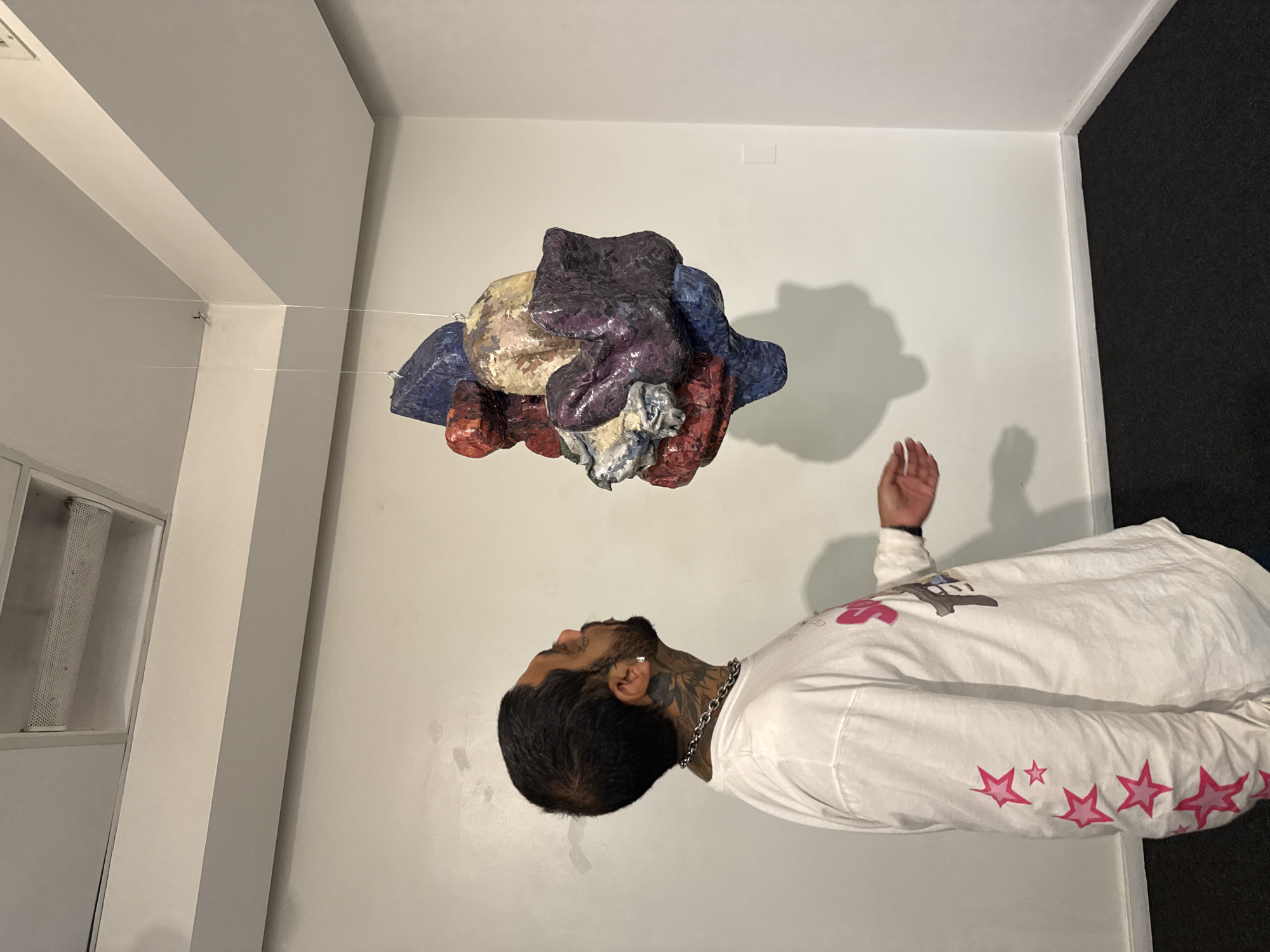 An indoor scene, likely an art gallery or studio in Buenos Aires, Argentina, captures a man observing a suspended abstract sculpture. The image depicts a man with dark hair, a beard, and visible neck tattoos, standing on the right side of the frame. He wears a white long-sleeved shirt adorned with pink star graphics on the sleeves and an indistinct graphic on the chest. A silver chain necklace and an earring are also visible. His right hand is slightly raised as he gazes upwards towards the artwork, suggesting contemplation or engagement.
Suspended from the white ceiling by thin, transparent wires, the multi-colored sculpture is the focal point. It is an abstract, textured piece composed of various crinkled or folded elements in shades of blue, purple, red, yellow, and off-white/light gray. The texture suggests a fragmented or mosaic-like surface. The sculpture casts a soft shadow on the white wall behind it.
The setting features plain white walls and a dark, carpeted floor on the right. A recessed light fixture is visible on the ceiling in the upper left corner. The overall lighting is even, suggesting an indoor environment with artificial illumination. The man's posture indicates focused attention or curiosity towards the unique artwork.
