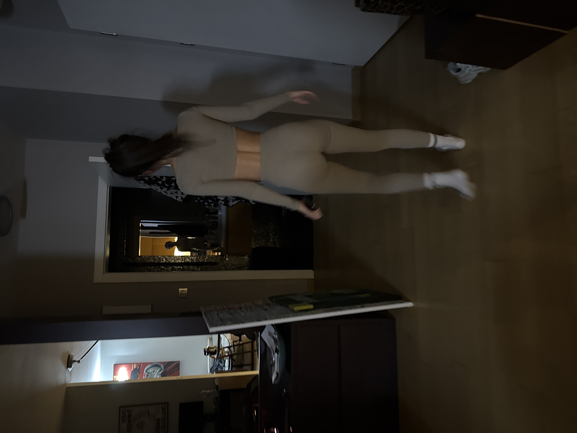 A woman in a beige athletic outfit stands in an interior space, likely a home or apartment in Warsaw, Poland. She faces away from the camera, her body angled slightly towards the left. The scene includes a hallway leading to another room, furniture (likely cabinets or a sideboard), a doorway, and interior design elements. The environment suggests a contemporary and somewhat dimly lit interior setting.