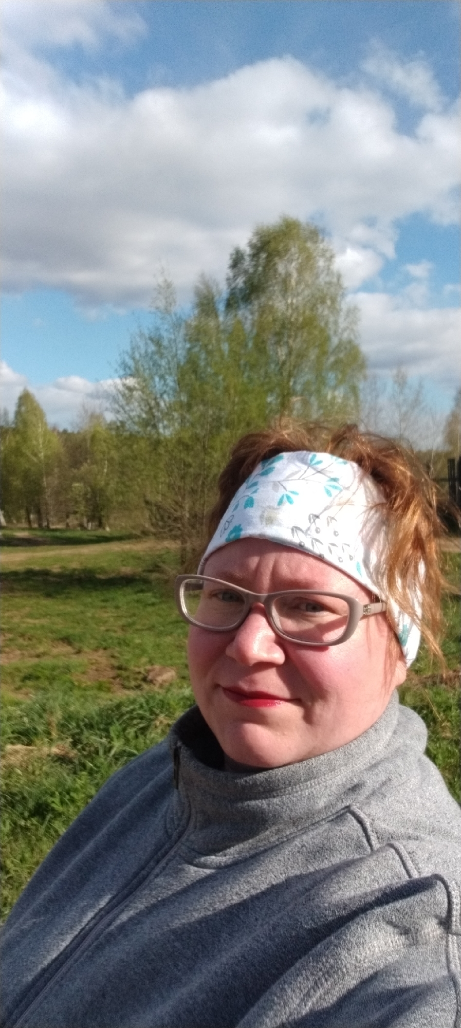 The image shows a woman in Зипуново, Russia, outdoors on a partly cloudy day. She is wearing a gray fleece jacket, a white headband with a blue floral pattern, and eyeglasses. She is facing the camera with trees and grass in the background.