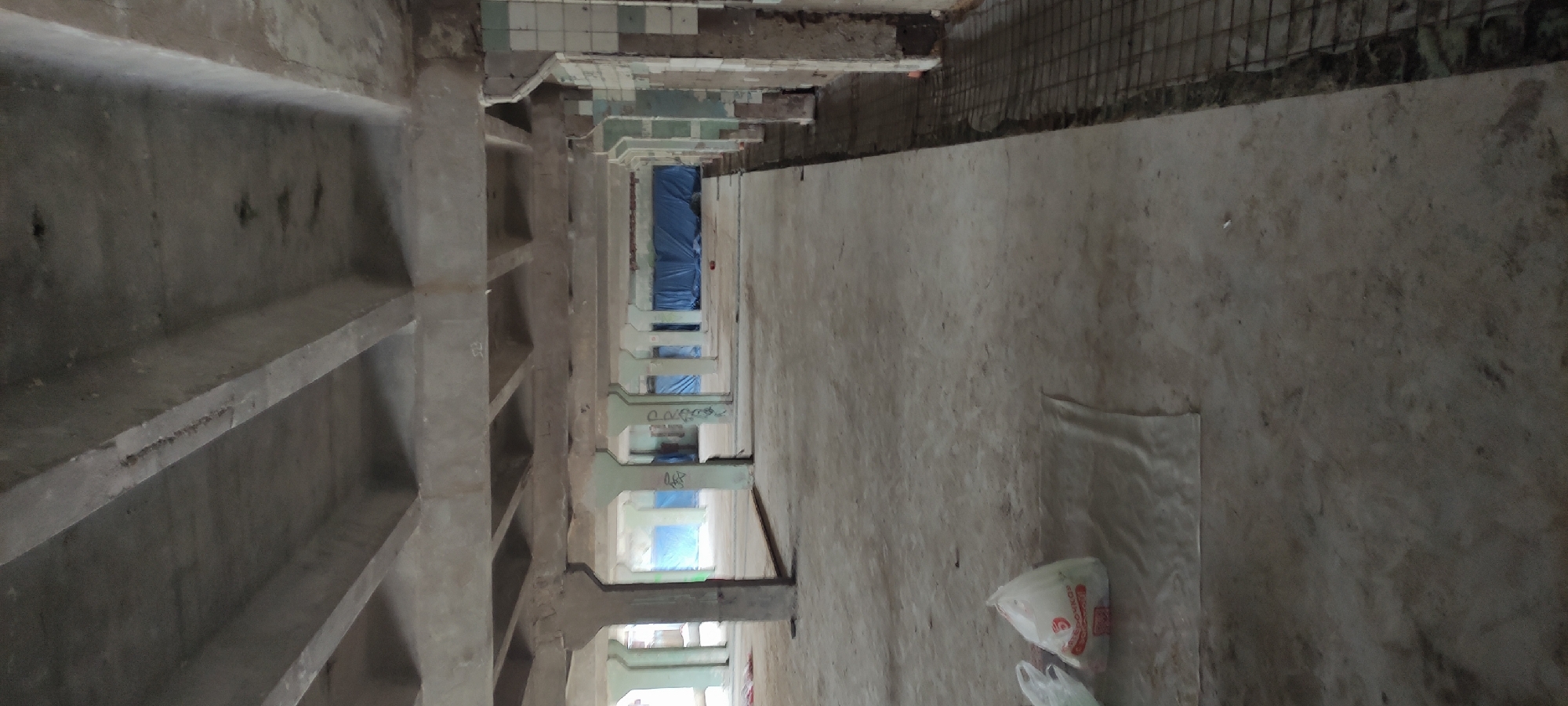 The image shows an interior perspective of a derelict or industrial structure, possibly located in the city of Fryazino, Russia. The view is taken looking down a corridor flanked by concrete support beams on the left side, which create a series of shelf-like recesses. These beams are in a state of disrepair, with visible signs of wear and age.
On the right side of the corridor, there is a concrete wall, also exhibiting signs of degradation. Debris, including white plastic bags, are scattered on the ground. The corridor extends into the distance, with the end partly obscured by what appears to be blue tarpaulin or sheeting. Above the visible portion of the corridor, there are sections of tile and exposed rebar. There are no people visible in the image. The overall environment is dark, suggesting poor lighting conditions, and exudes an atmosphere of abandonment or neglect.
