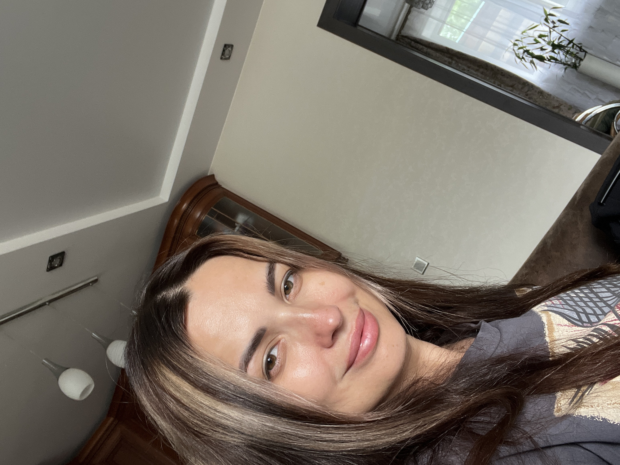 A woman is taking a selfie indoors. She has long, brown hair with blonde highlights and brown eyes. She is wearing a dark gray t-shirt. The background includes a ceiling with lights, a wooden cabinet, a wall, a door with a window, and a plant in a white pot. The scene appears to be in a residential setting.