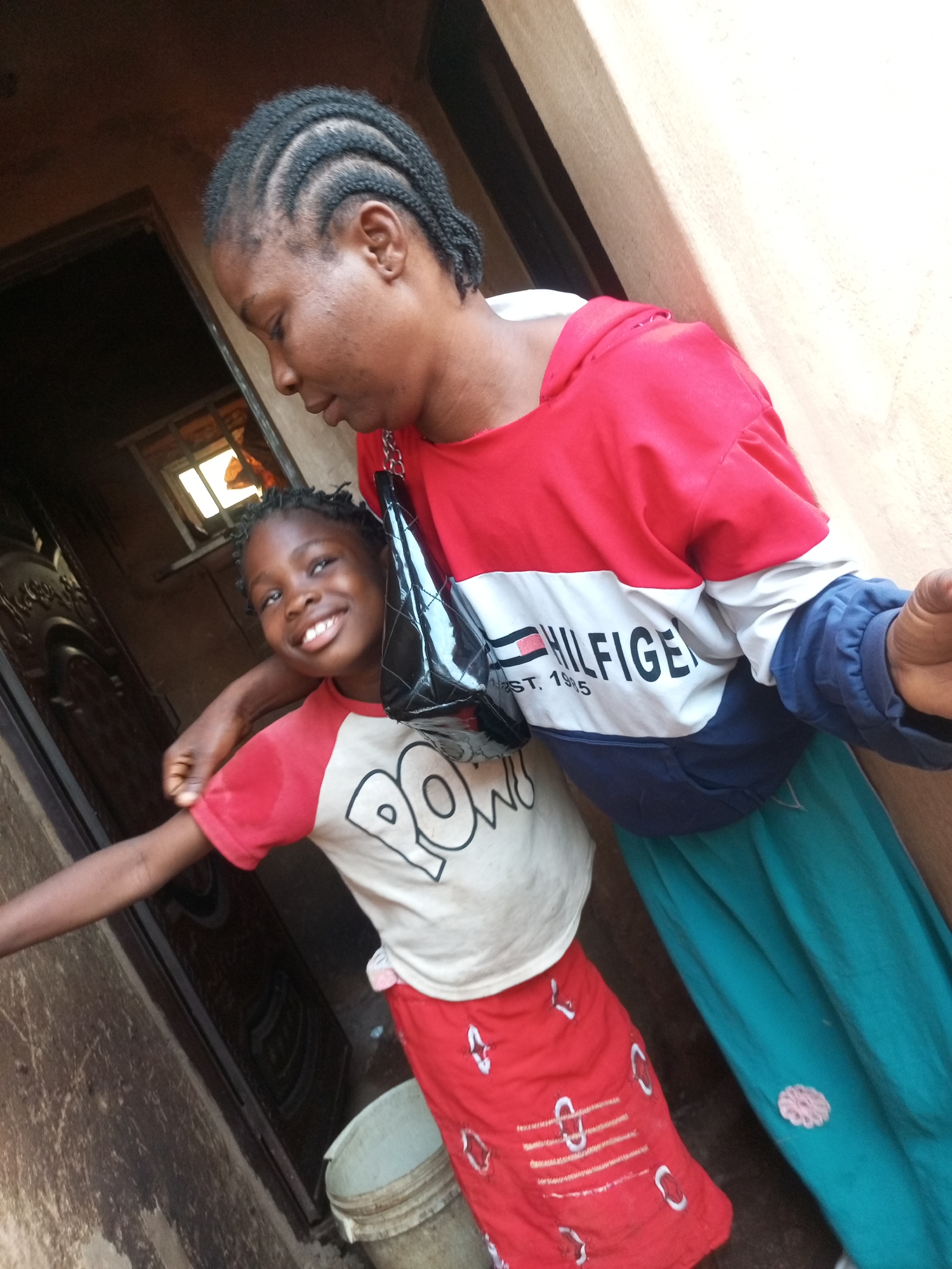 The image depicts a mother and daughter posing for a photo outdoors, possibly in a residential area of Tempure, Nigeria. The mother, on the right, is wearing a red, white, and blue Tommy Hilfiger sweatshirt and a teal skirt. She has braided hair and is holding a black purse. The daughter, on the left, is wearing a red and white t-shirt with "ROW" printed on it and a red skirt with white accents. She is smiling and has her arm around her mother. They are standing in front of a textured wall and a partially visible doorway. The lighting suggests it is daytime.