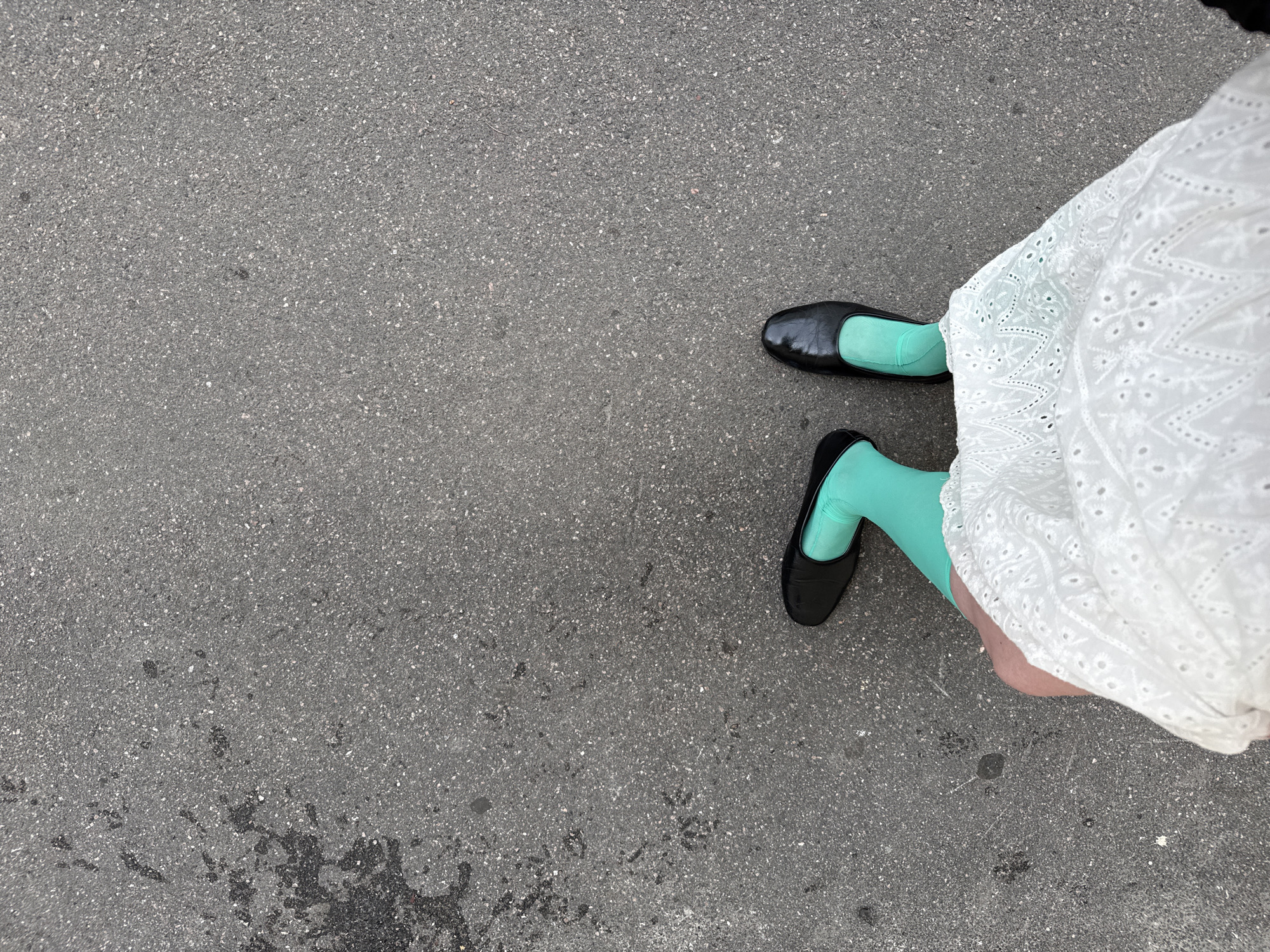 The image is an overhead, ground-level shot of a person's legs and feet standing on asphalt. The person is wearing black flat shoes, bright turquoise hosiery, and a white eyelet lace skirt. The asphalt appears to be a public walkway, possibly a sidewalk or pedestrian area, with some dark stains, possibly oil or water, on the left side of the frame. The lighting suggests it is daytime. This scene is not specifically identifiable as being in Saint Petersburg, Russia, as it lacks any distinctive landmarks or cultural markers.