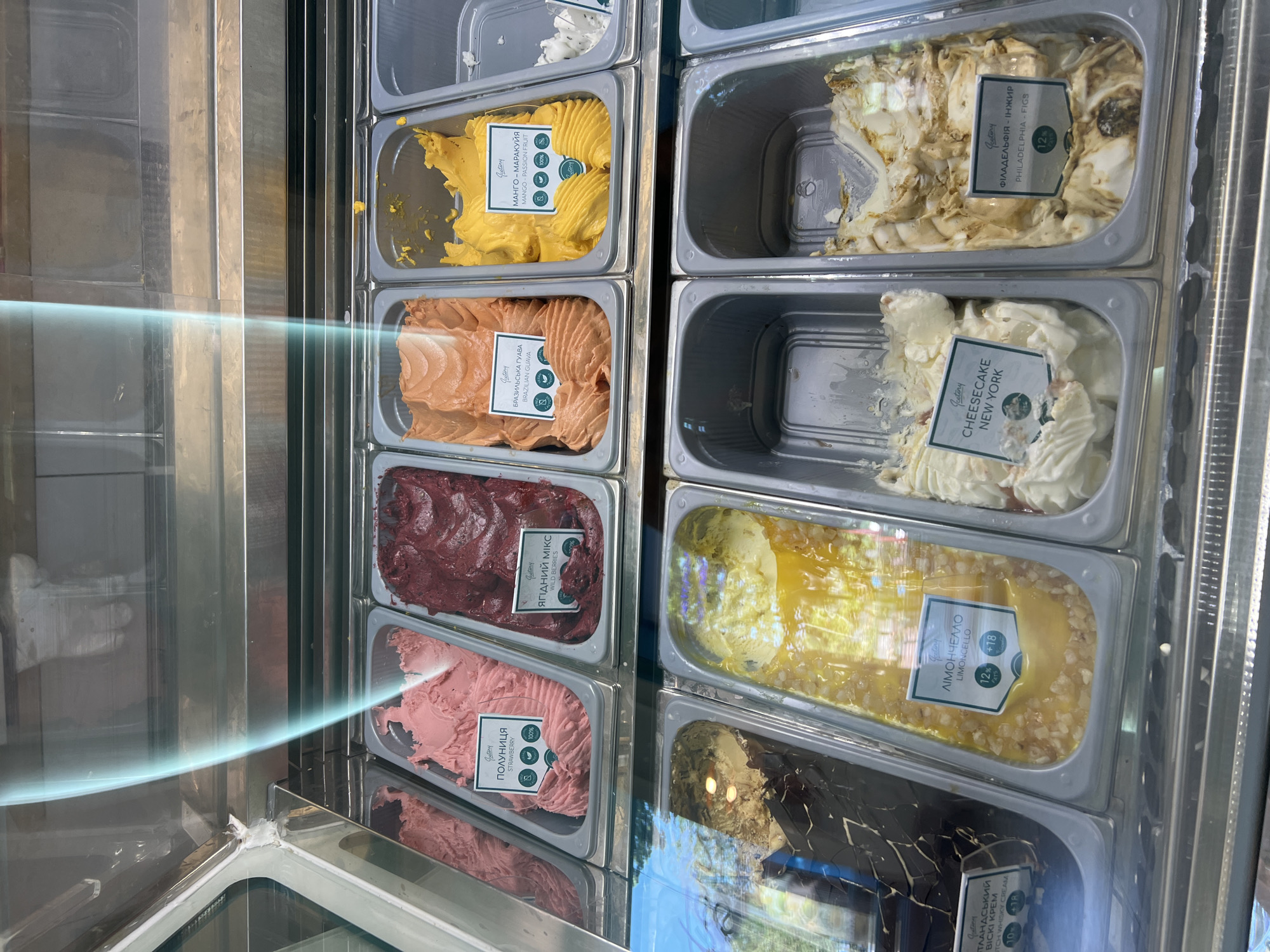 This is a close-up, eye-level shot of an ice cream display case at an establishment in Odesa, Ukraine. The display is well-lit and filled with various flavors of gelato in metal tubs. The tubs are arranged in two rows.
In the top row, from left to right, are: a pink strawberry flavor, a dark red berry flavor, an orange Brazilian guava flavor, and a vibrant yellow mango-passion fruit flavor. Labels are visible for the strawberry, Brazilian guava, and mango-passion fruit.
The bottom row, from left to right, shows: a dark chocolate and cream swirl flavor, a yellow limoncello flavor with nuts, a cheesecake new york flavor, and a philadelphia-figs flavor. Labels are visible for limoncello, cheesecake new york, and philadelphia-figs.
The overall environment appears clean and professional, typical of an ice cream shop. Reflections on the glass display case show what appears to be the interior of the shop, including metallic shelving. No people are directly visible in the frame, but a hand is faintly reflected in the upper left corner of the display case glass, possibly indicating an employee or customer interacting with the display. The lighting creates a slight glare and highlights the textures of the ice cream.