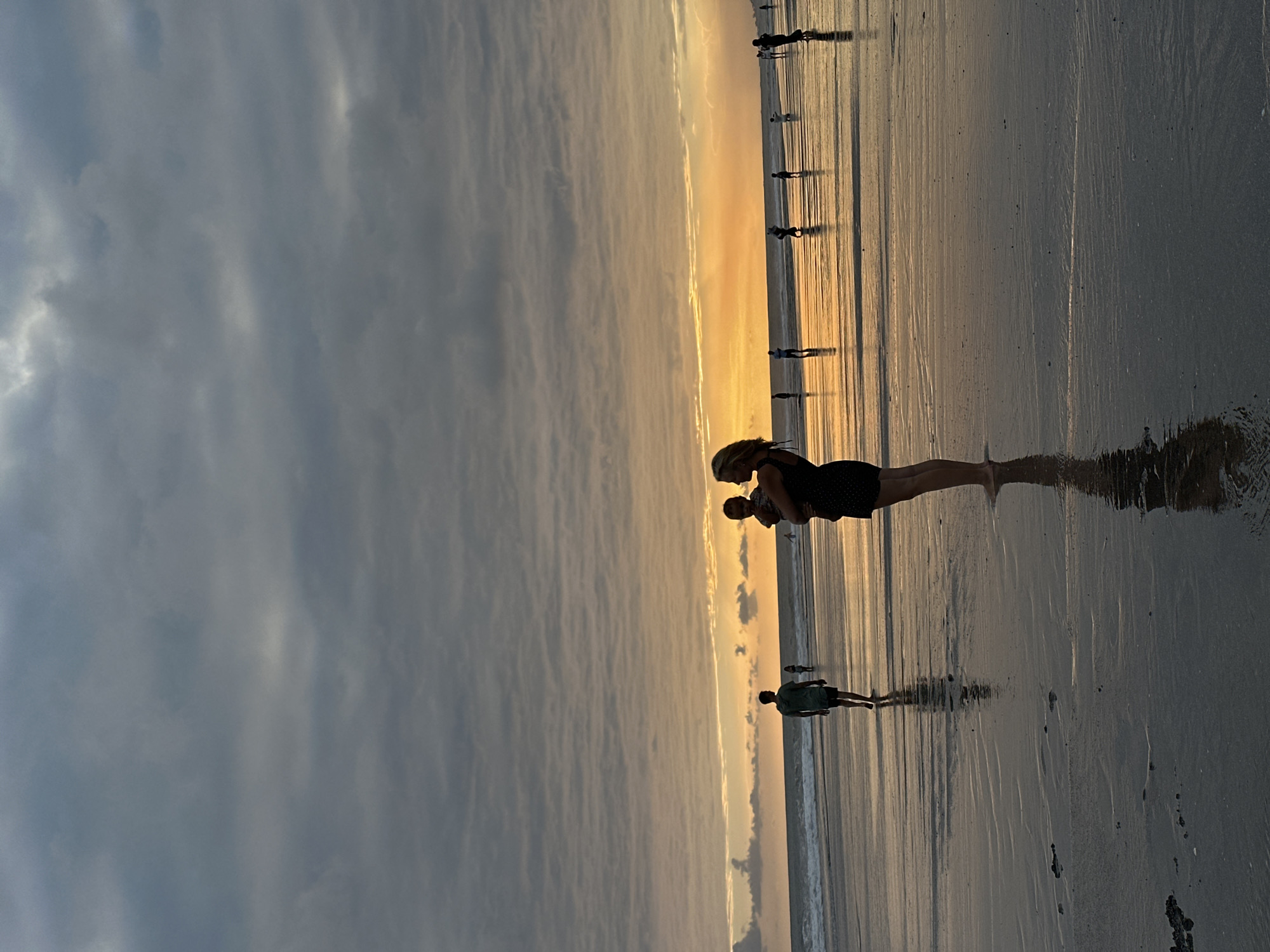A silhouette of a woman holding a baby is in the foreground, facing left, at the edge of the water on a beach at sunset. The woman has blonde hair and is wearing a dark dress. The baby is facing the woman and is wearing light-colored clothing. The wet sand reflects the sky and the figures. In the mid-ground, other figures are walking along the beach, and the ocean is visible with waves. The background shows a cloudy sky with a bright orange sunset. This scene is taking place on the beach in North Kuta, Indonesia.