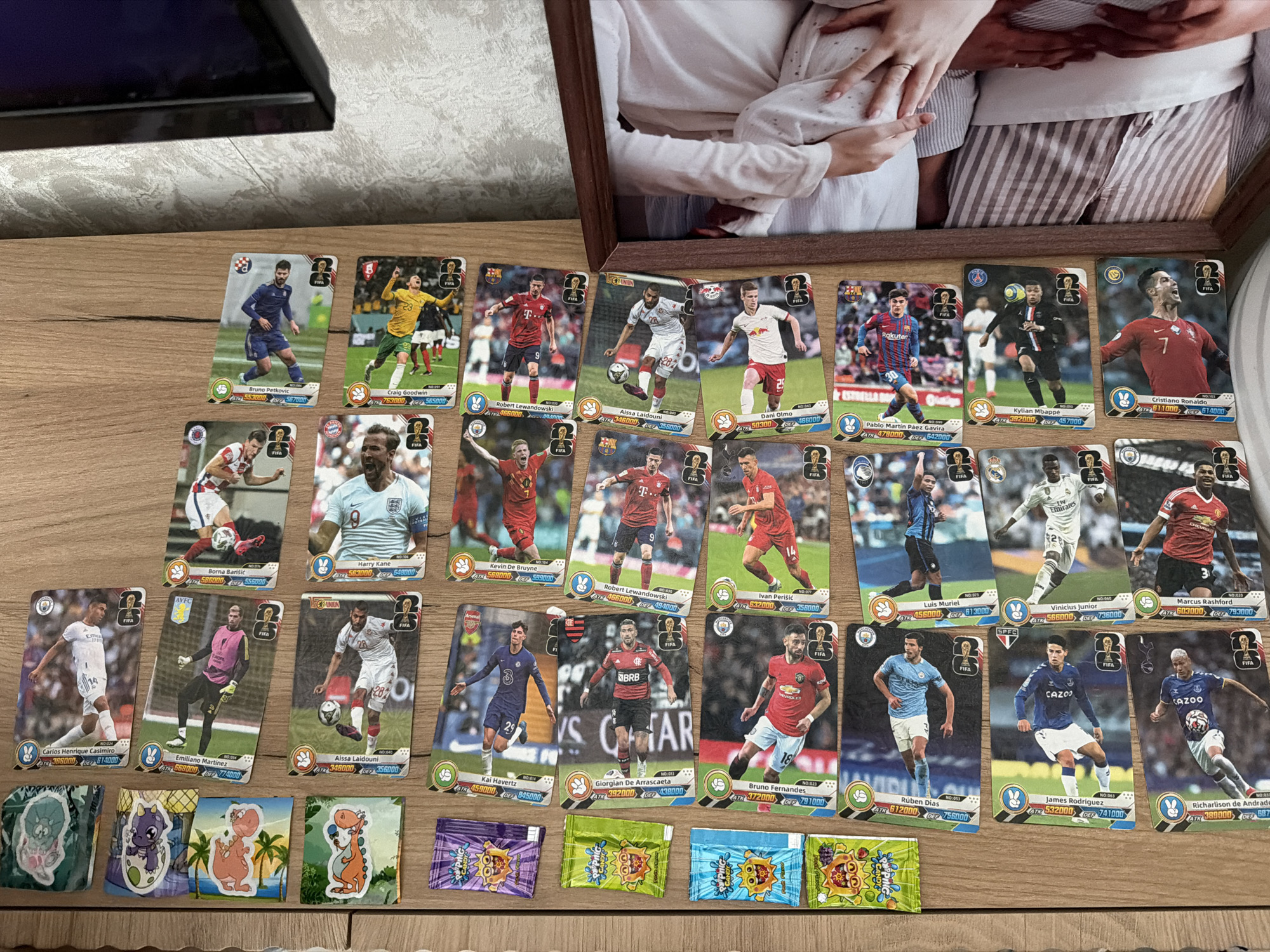 The image displays a collection of football (soccer) trading cards arranged on a wooden surface, possibly a table. The cards feature images of various football players in action, along with their names, team affiliations, and statistics. Some of the players visible include Kai Havertz, Robert Lewandowski, and Cristiano Ronaldo. In addition to the football cards, there are also several brightly colored candy wrappers scattered among them, specifically "Popping Candy" wrappers with cartoonish characters. To the right of the wooden surface, a framed photograph is partially visible, showing a person's arms and torso, suggesting a personal touch or display. The background of the image is a textured wall, possibly wallpapered, with a section of what appears to be a television screen visible in the upper right corner, partially obscured by the framing of the photograph. The scene does not provide any specific indicators of its location in the city of Kazan, Russia.