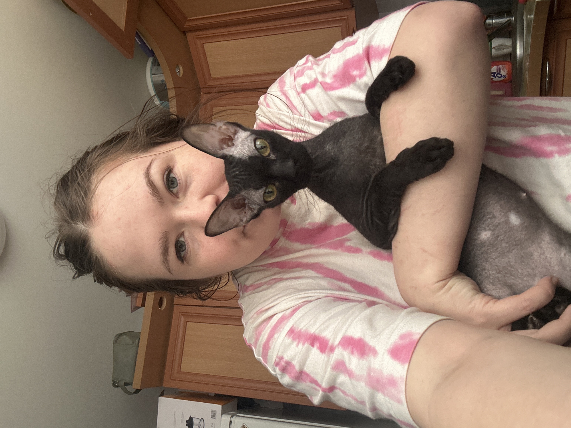 A woman is holding a gray cat with black paws and yellow eyes, close to her face. The woman is fair-skinned and wearing a pink and white tie-dye shirt. The background features light wood cabinets and a wall painted in a light color. Based on the context of the image, it's likely taken in an indoor environment, possibly a kitchen. The location is unknown.