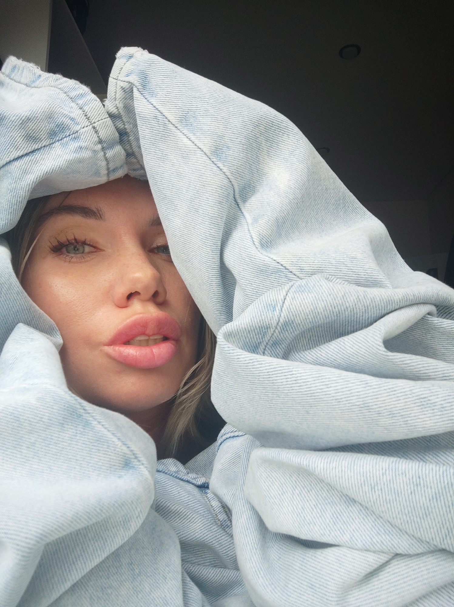 A fair-skinned woman with blonde hair and green eyes is positioned in the foreground. Her face is partially framed and obscured by light blue denim material. Her lips are pursed in a pouting expression. The environment behind her is dark, only revealing a ceiling with a recessed light fixture. The location is not discernible as Votkinsk, Russia from the information in the image.
