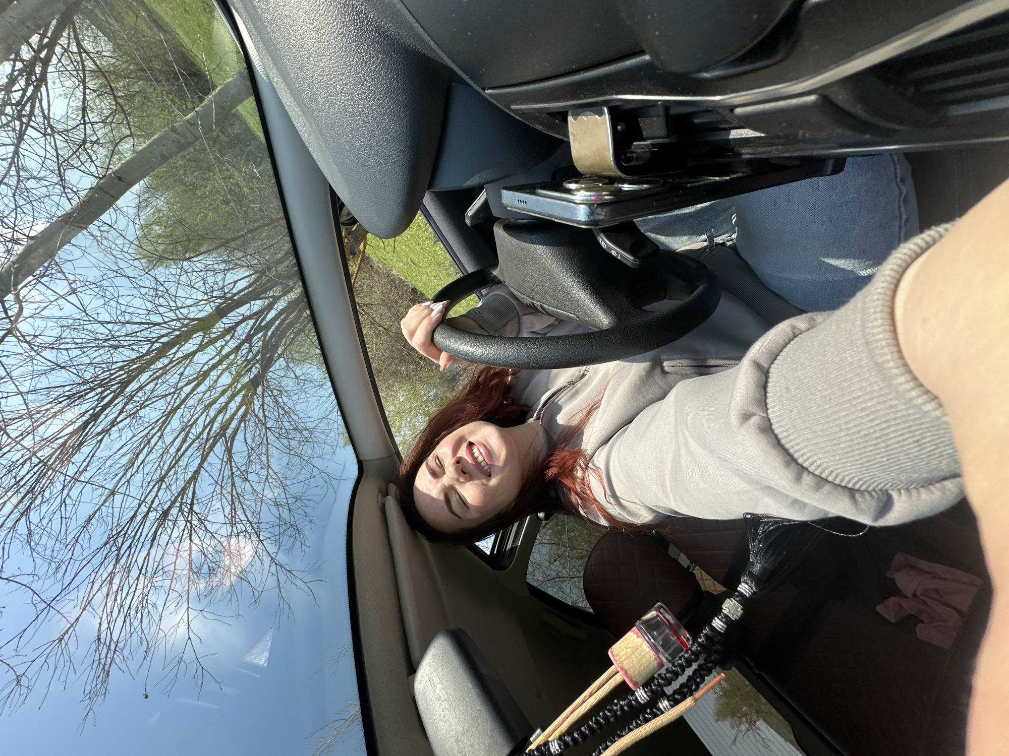 A young woman with reddish-brown hair and fair skin is taking a selfie inside a car. She is in the driver's seat, holding the steering wheel and smiling at the camera. She is wearing a gray hooded sweatshirt and blue jeans. The car's interior is visible, including the dashboard and windshield. Outside, trees and a blue sky are seen through the windshield, potentially indicative of an outdoor setting. There is no definitive information to confirm the location as the city of Cheryomushki, Russia. Other objects, such as a car phone holder, are visible as well as what appears to be brushes or decorative items.