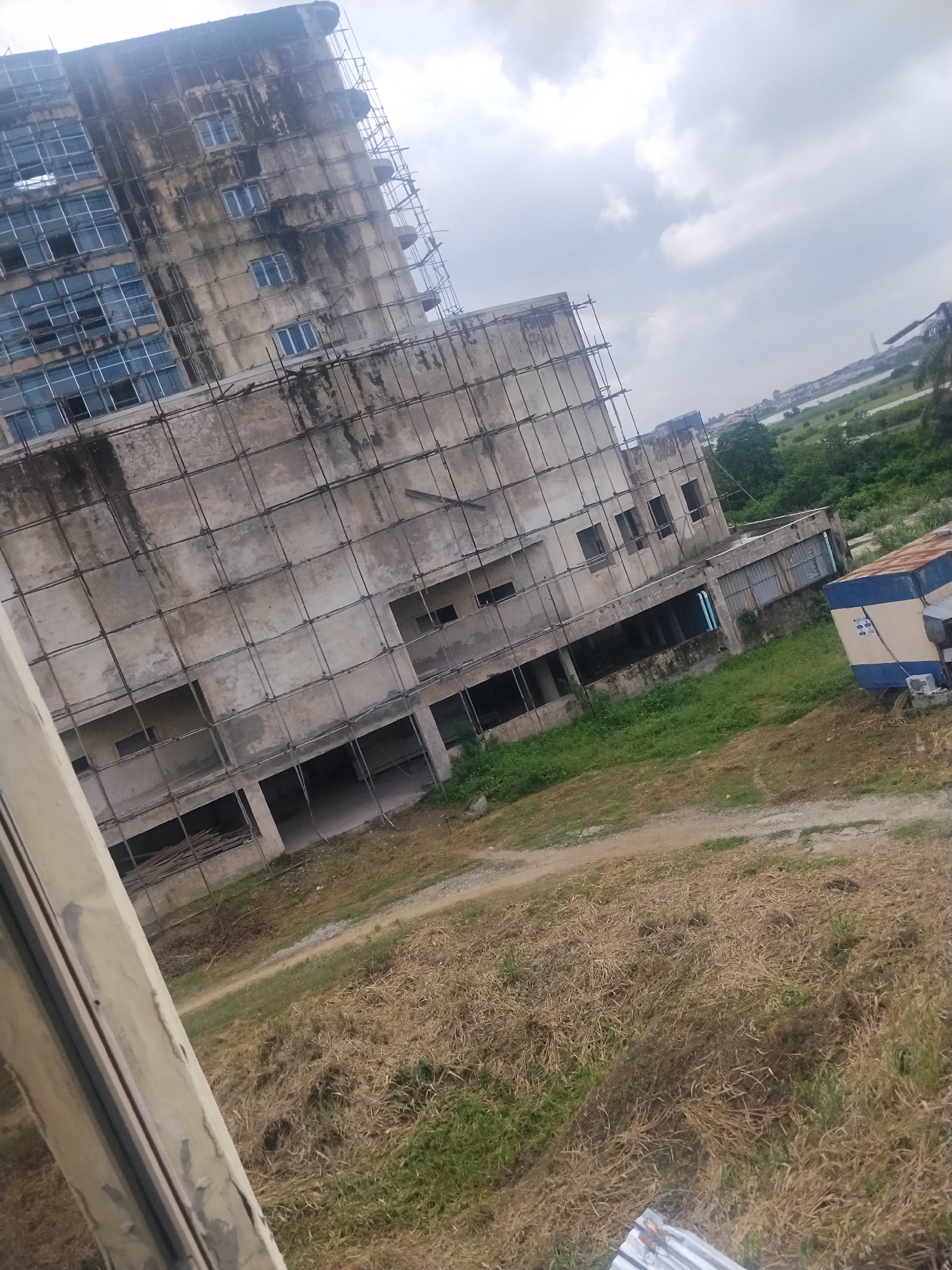 The image captures an unfinished multi-story building, possibly a hotel or office complex, in Lagos, Nigeria. The building, with its weathered concrete facade and scaffolding, appears to be under construction or renovation. The windows are largely intact, some showing reflective glass. The surrounding environment is characterized by dry, patchy grass, a dirt road, and lush green vegetation in the background. In the distance, a body of water, likely the Lagos Lagoon or the Atlantic Ocean, is visible, with the hint of a bridge and other structures. A small, blue and yellow utility building is situated to the right of the main structure. The sky is overcast with scattered clouds, suggesting a typical Lagos day. The scene is devoid of any visible people or human activity, creating a sense of stillness and neglect.