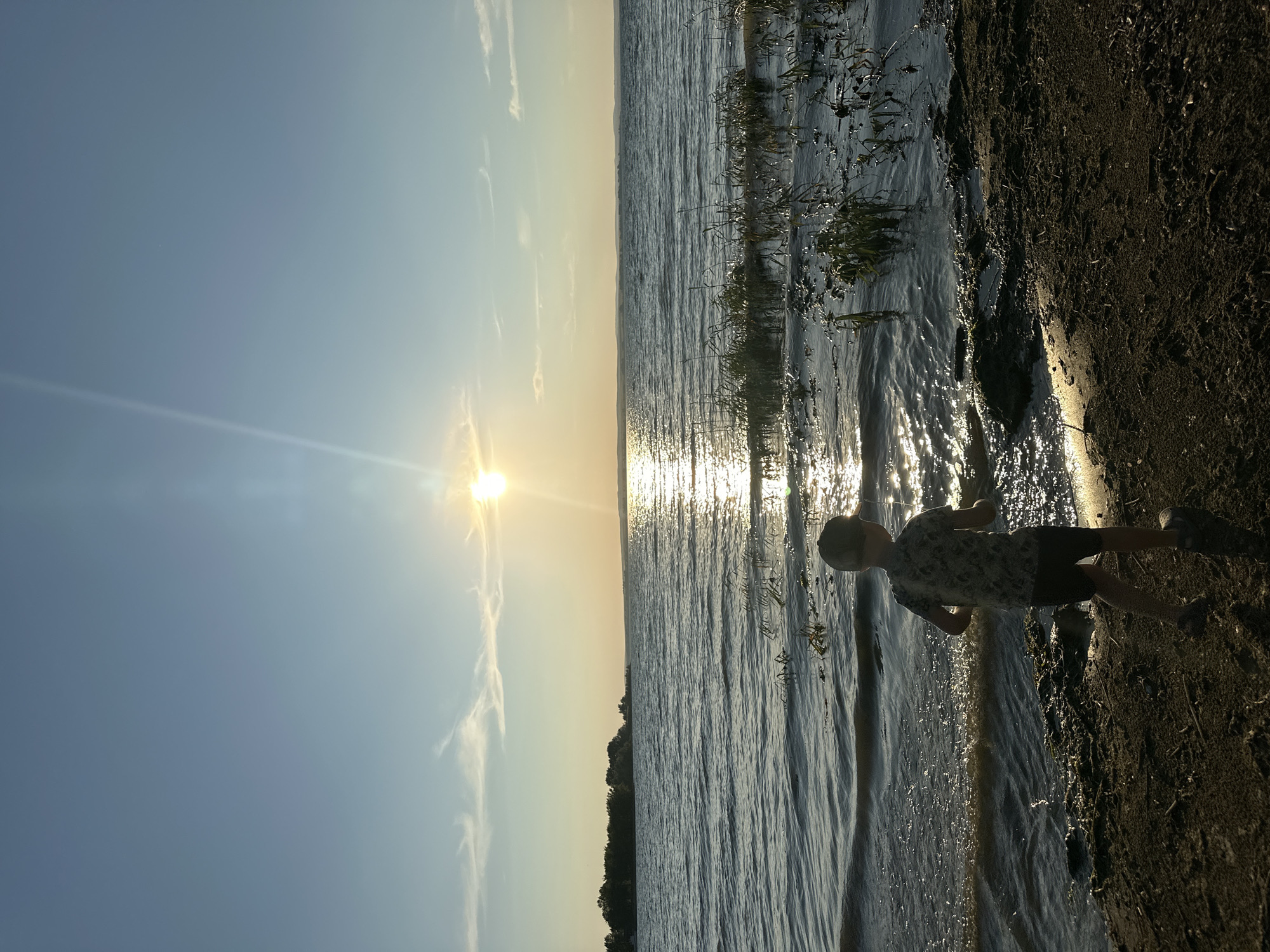 A lone young boy, wearing a patterned t-shirt, dark shorts, and a baseball cap, stands on a muddy shore, facing away from the camera. He appears to be looking out at a calm body of water, possibly a lake or river, under a clear blue sky with a bright sun setting or rising. The sun's rays create a shimmering reflection on the water's surface. In the distance, a tree-lined shore is visible. The scene is set at the water's edge in Energetik, Russia, during what appears to be a warm day. The overall mood of the image is tranquil and contemplative.