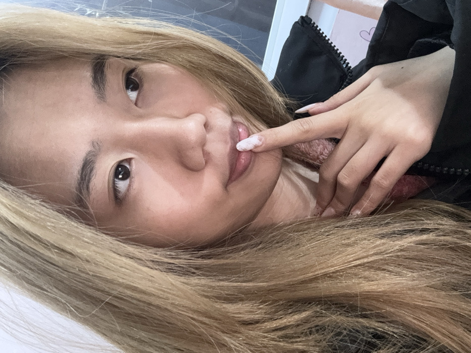 A close-up image depicts a young East Asian female, visible from approximately the chest up, in an indoor setting within Shapingba District, China. She has long, straight, blonde hair, dark eyes, and a light complexion. Her lips are slightly parted and appear glossy. She is dressed in a dark, possibly black, zip-up jacket worn over a light-colored garment, with a textured, light pink layer visible beneath. Her right hand is raised, with a long, manicured index finger, featuring a white marble-patterned nail, lightly contacting her lower lip. Her gaze is directed towards the upper right of the frame. The background is blurred, showing indistinct light and dark areas, including a partial white horizontal element and a faint pink object on the far right.