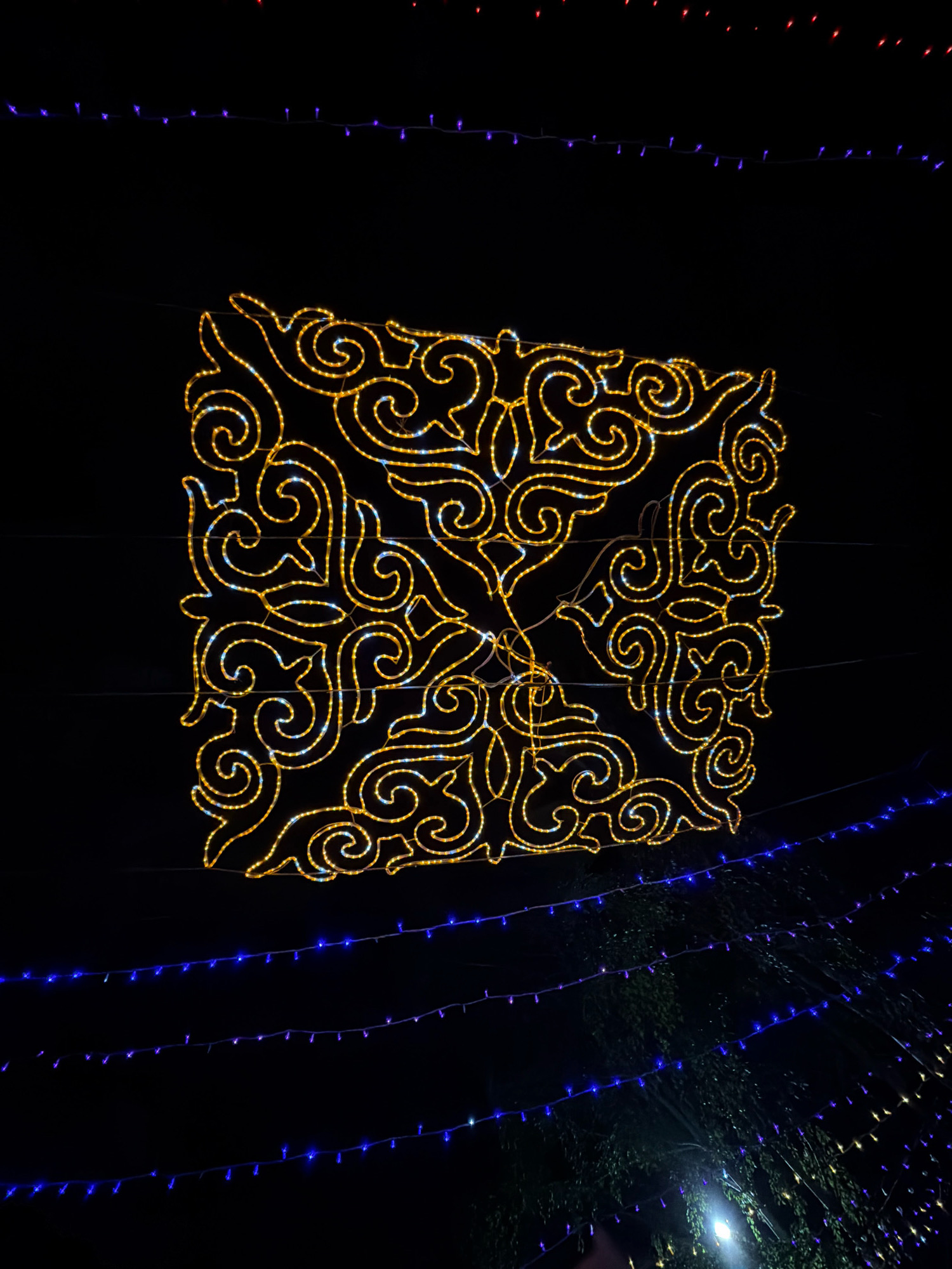 An ornate, square-shaped illuminated decoration, constructed from yellow and white lights, hangs against a dark night sky. The pattern within the square resembles intricate swirling motifs, possibly inspired by traditional Kazakh designs. Below the central decoration, several strings of blue and purple LED lights are stretched horizontally, creating a layered effect. In the lower right portion of the image, the branches and leaves of trees are faintly visible, illuminated by a bright, out-of-focus light source. The overall scene suggests an outdoor nighttime display, likely in a public area decorated for an event or holiday. The specific location in Pavlodar, Kazakhstan, is unknown from the image alone, but the presence of such decorative lighting is common in city centers and parks during festive periods.