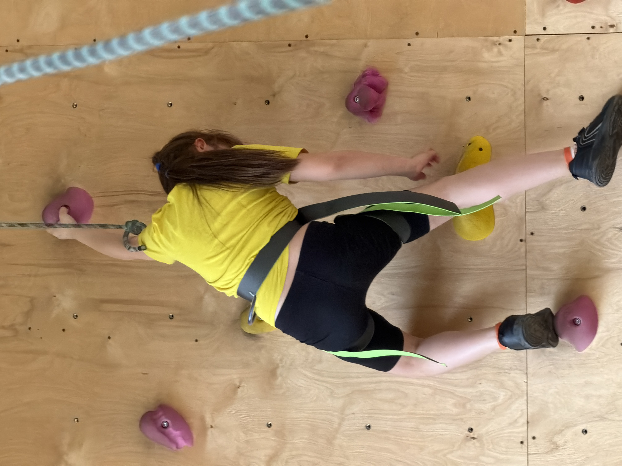 A person is captured in a dynamic pose while rock climbing indoors in Izhevsk, Russia. The subject, likely a child or young individual, is seen from an overhead perspective, positioned horizontally across a light-colored plywood climbing wall. They have long brown hair, wear a bright yellow short-sleeved t-shirt, black shorts, and black climbing shoes with orange accents. A grey and neon green climbing harness is visible around their waist and thighs, connected to a light-colored climbing rope that runs diagonally across the upper left of the frame.
The climber's arms are extended, with the left hand gripping a pink climbing hold. Their legs are also stretched out, with the right foot planted on another pink hold towards the lower right. A yellow climbing hold is visible near the person's right hand and another pink hold is seen above their head. The plywood wall features numerous small bolts, indicating where holds can be attached. The scene depicts an active and engaged climbing session, emphasizing the stretch and balance required for the sport. There is no visible text in the image.