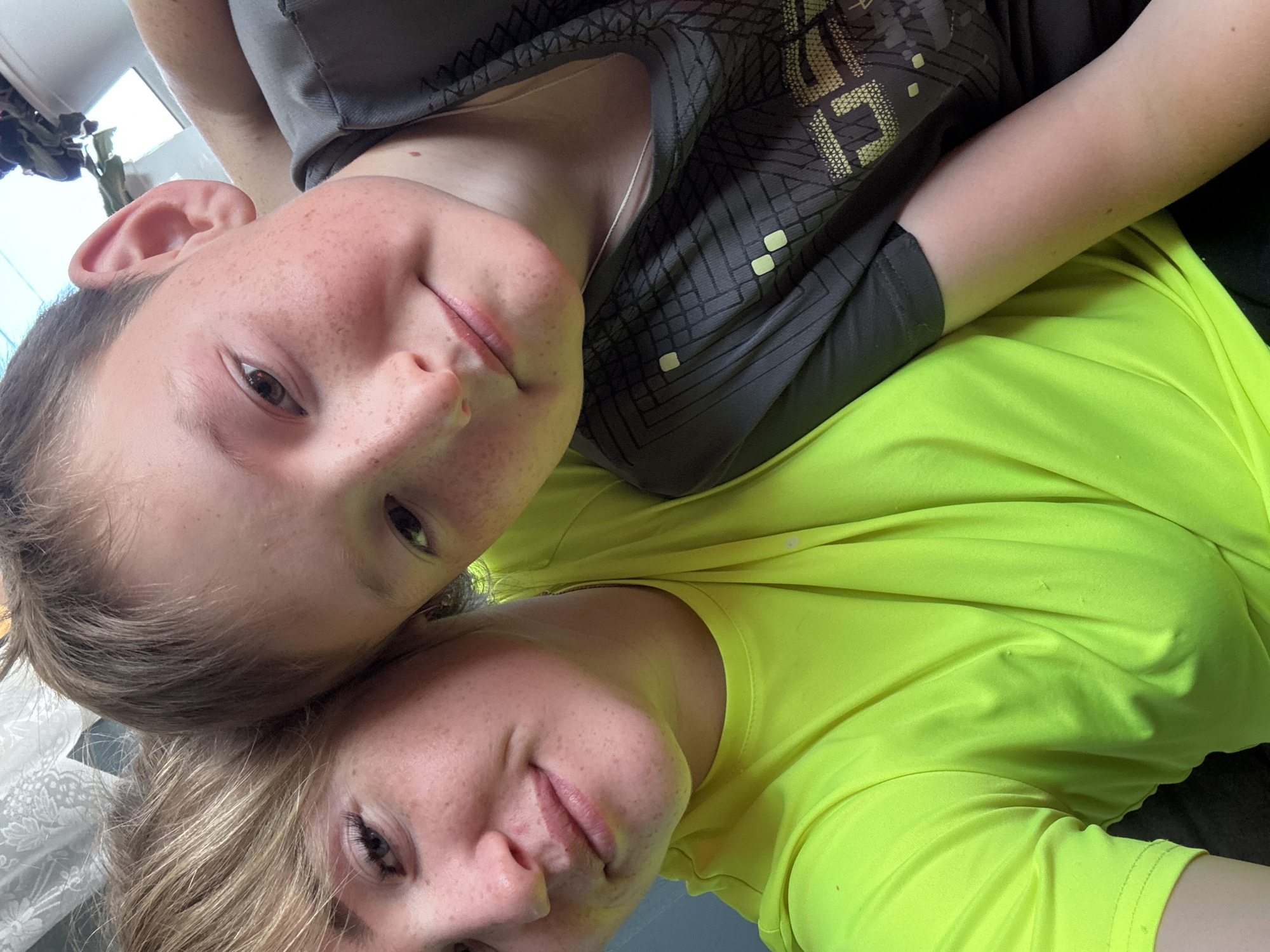 This image, taken in the city of Ляльшур, Russia, displays a close-up, selfie-style photograph of two individuals indoors. On the left, an adult female with light-colored hair, noticeable eyelashes, and freckles on her face is visible, wearing a vivid lime-green short-sleeved shirt. A light-colored strap or lanyard is partially visible around her neck. On the right, a younger male, likely a child or teenager, with light brown hair and prominent freckles across his nose and cheeks, looks towards the camera with a neutral expression. He is wearing a dark grey short-sleeved shirt featuring a geometric pattern in lighter shades, and a thin white chain necklace. The two individuals are positioned closely together, suggesting a familiar relationship. The background reveals an indoor setting with a white, possibly lace, curtain in the upper left, and a dark leafy plant in a white pot against a bright, possibly sunlit, background in the upper right.