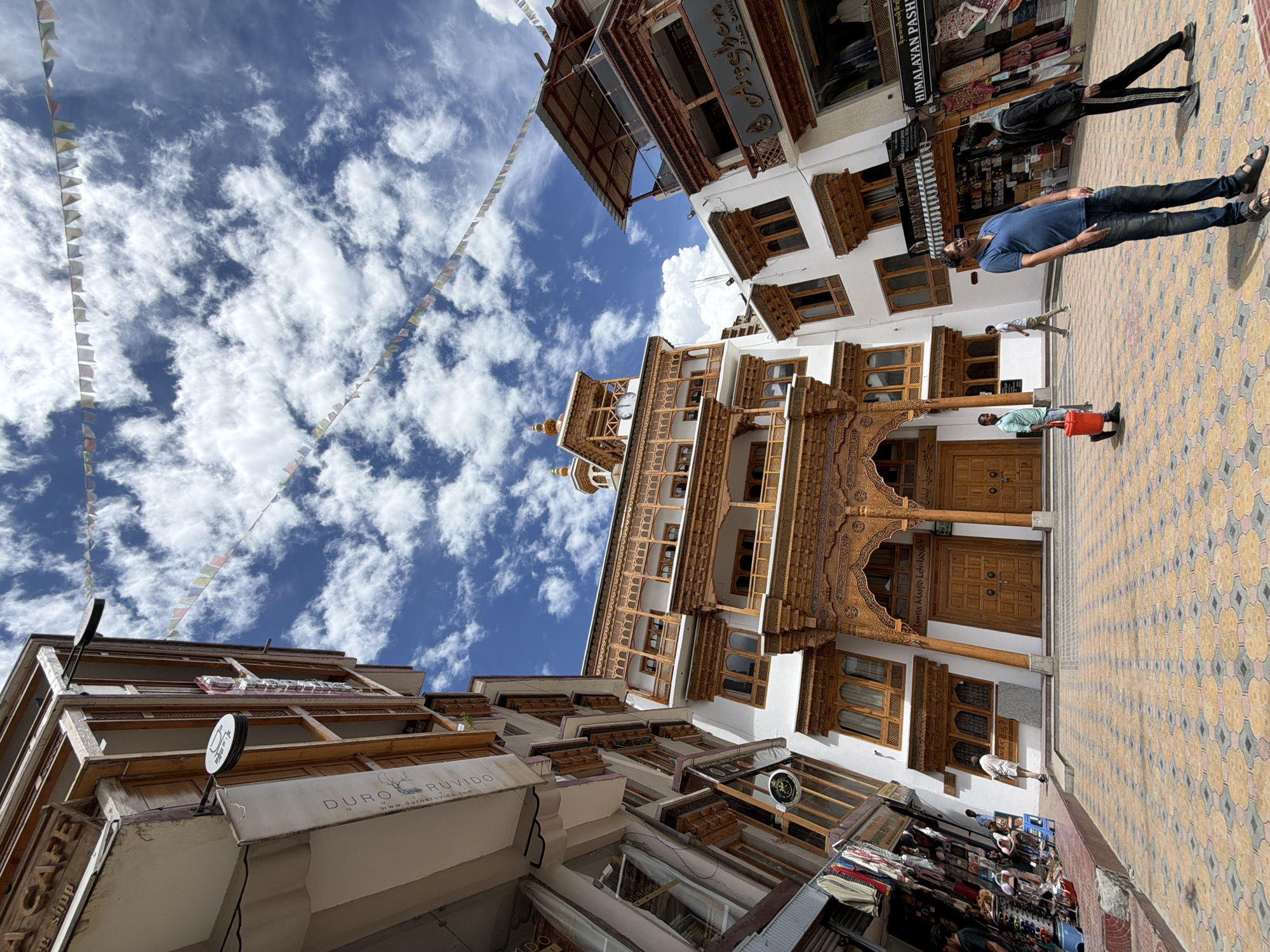 The image captures a street scene in Leh, India, likely near the Jama Masjid. The architecture is characterized by traditional Tibetan style, featuring white facades, wooden balconies with intricate carvings, and ornate window frames. Prayer flags are strung across the sky, which is a vibrant blue dotted with white clouds.
In the foreground, a man in a blue t-shirt and jeans stands facing to the left, his body tilted. To his right, another man, wearing a black tracksuit, is seen from the back, also looking towards the left. Further left, a man in a light blue shirt walks with a red bucket. In the background, several other individuals are visible, some interacting with shops lining the street.
The ground is paved with decorative tiles in a geometric pattern. Shops are visible on either side of the street, with displays of goods, including textiles and clothing. The overall atmosphere suggests a bustling day in a tourist-oriented area. The presence of "Jama Masjid Leh-Ladakh" signage on the building on the right confirms the location.