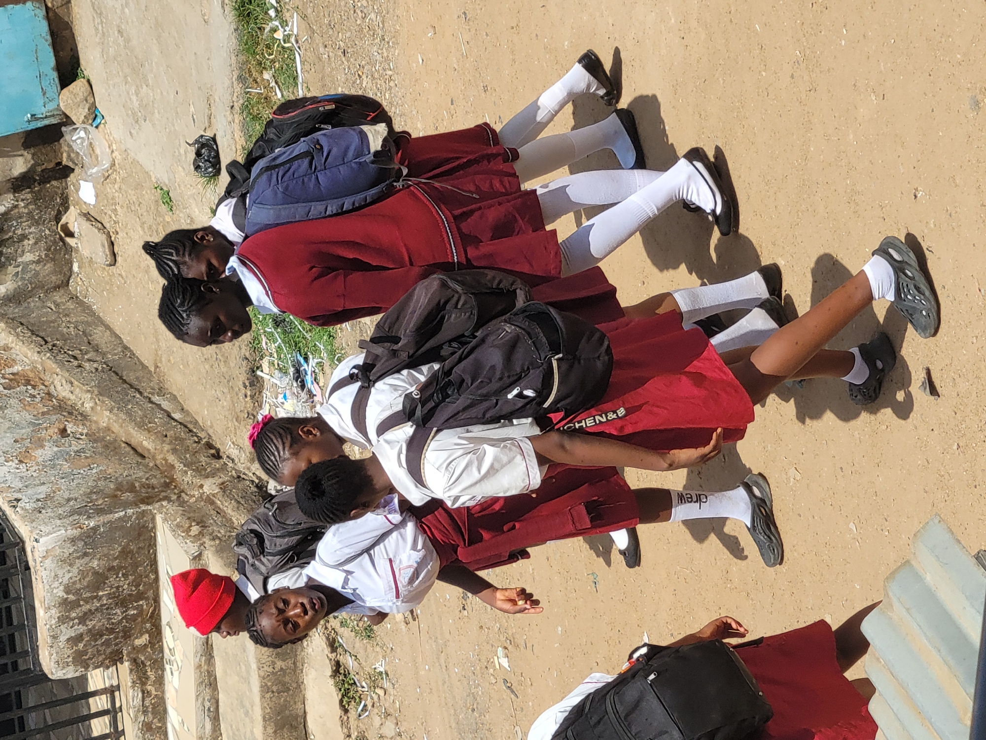 Four young girls wearing school uniforms consisting of white shirts, maroon skirts, and white socks are positioned closely together outdoors in Jos, Nigeria. They each wear a backpack. The environment appears to be a dirt road or yard, with evidence of litter. A building or structure, possibly with steps, is visible to the left. The girls are either standing or sitting close together. One girl wears a red beanie.