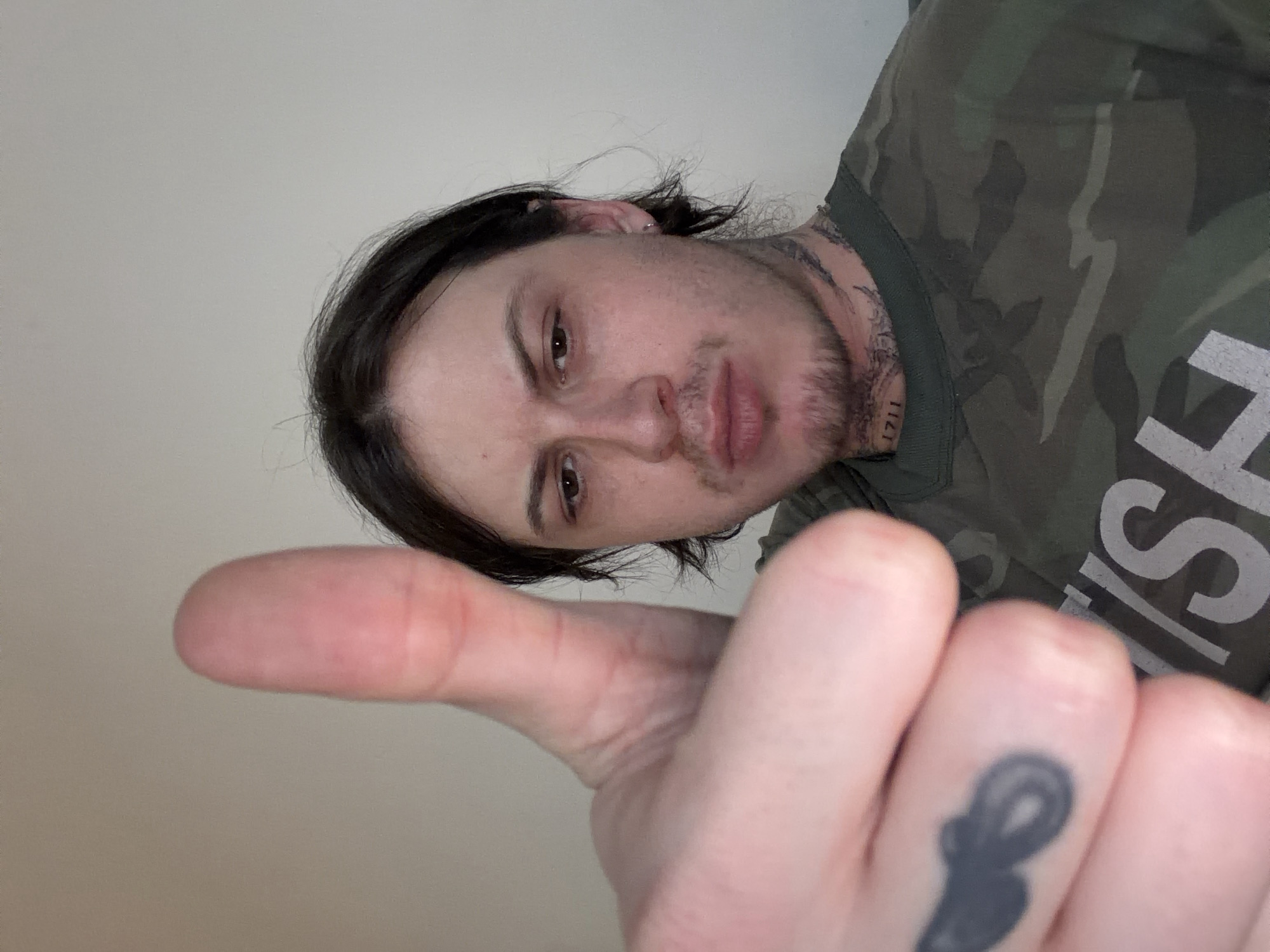 A person with dark hair and a slight beard is giving a thumbs-up. The person is wearing a camouflage t-shirt. A tattoo is visible on their finger and neck. The background is a plain white wall. There is no indication of the location in London, United Kingdom.