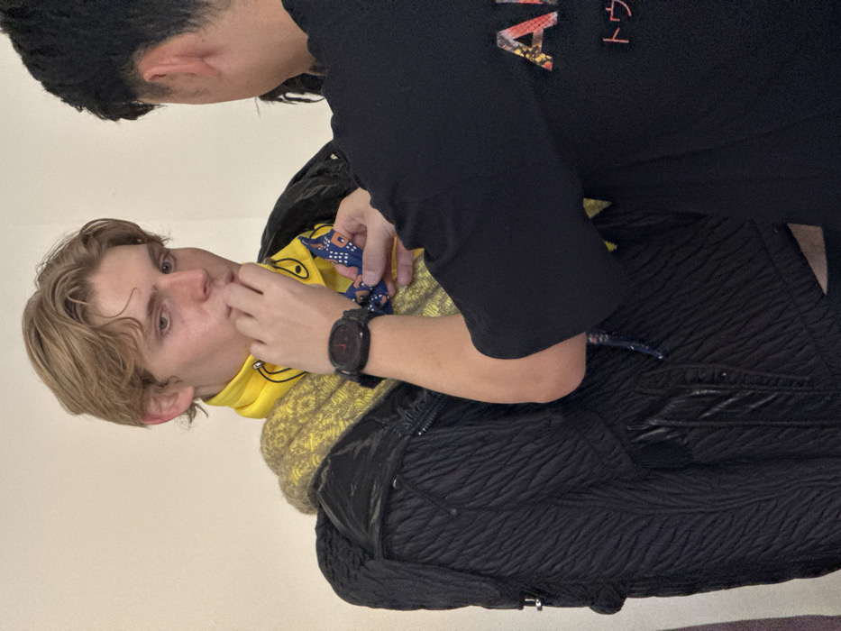 An indoor scene in Milan, Italy, captures two individuals engaged in a styling or fitting process against a plain, light-colored wall. On the left, a fair-skinned male with blonde hair, visible from the chest up, looks downwards, with his right hand touching his mouth. He is dressed in layered clothing: a yellow neck scarf with black cartoonish face outlines, an underlying textured yellow-green knit garment, and a prominent black crinkled or quilted jacket. To his right, partially obscured and seen from the back, is a second individual wearing a black t-shirt displaying partial text, including "AI" (or "AR") in a textured pattern and Japanese characters. This individual's right hand, extending from a sleeve, is manipulating a small, dark blue patterned fabric accessory near the first individual's neck, while their left wrist is adorned with a black smartwatch featuring a red display element.
