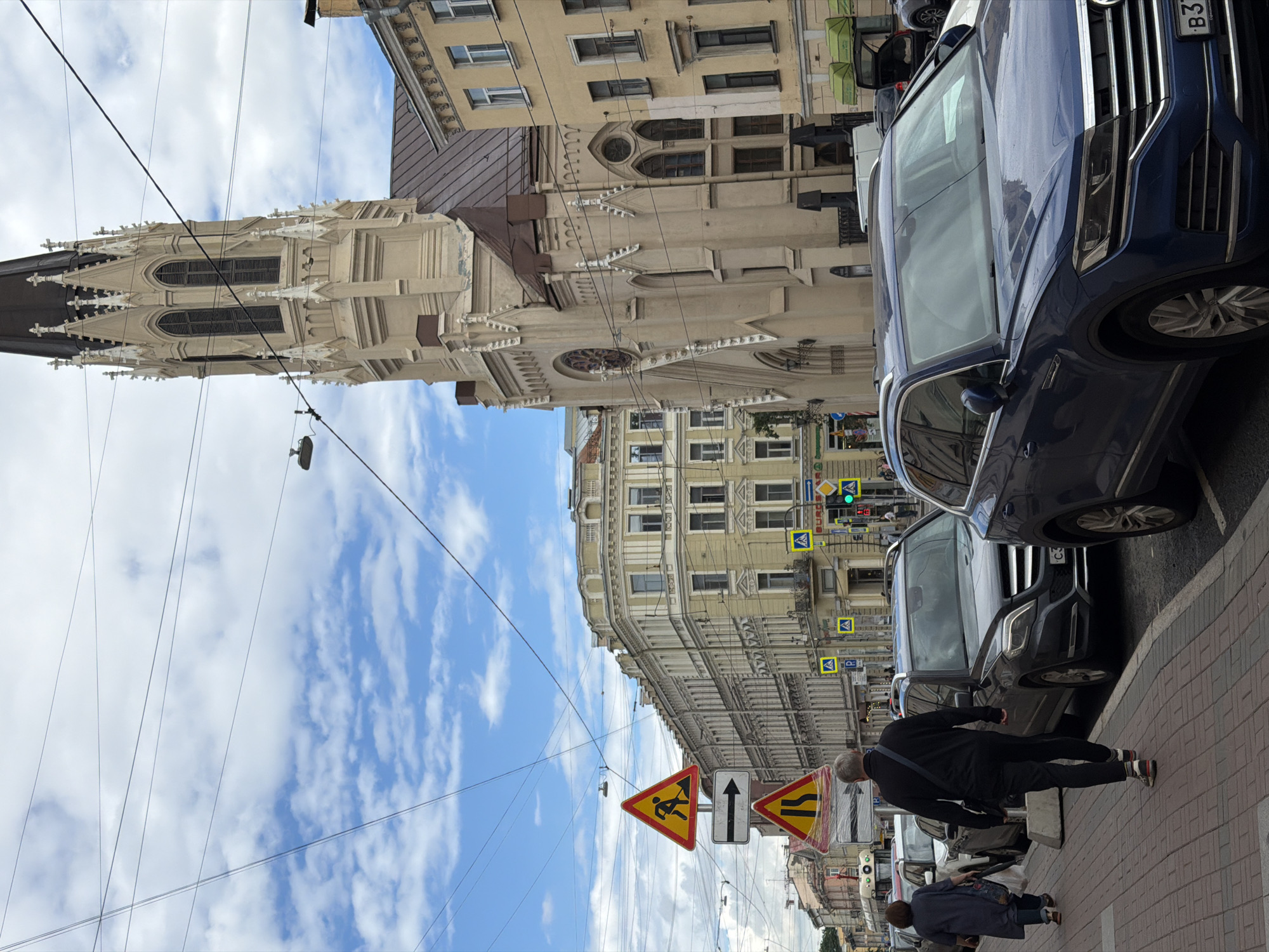 The image depicts a street scene in Saint Petersburg, Russia, likely during the daytime with partly cloudy weather. Several buildings line the street, showcasing architectural styles that suggest an older European city. One prominent building appears to be a historical church or cathedral with gothic architectural elements, including pointed spires and ornate detailing. Adjacent to it, other buildings exhibit a more classical facade with numerous windows and decorative cornices.
On the street, parked cars are visible, including a blue SUV in the foreground. The pavement is made of brick. Several traffic signs are present, some indicating road work and pedestrian crossings. A few individuals are on the sidewalk. One person, a man with graying hair, is walking away from the camera. Another person, a woman, is also walking on the sidewalk. Overhead, a network of electrical wires crisscrosses the sky. The overall atmosphere appears to be that of a bustling urban environment with historical architecture. The sky is blue with scattered white clouds.
