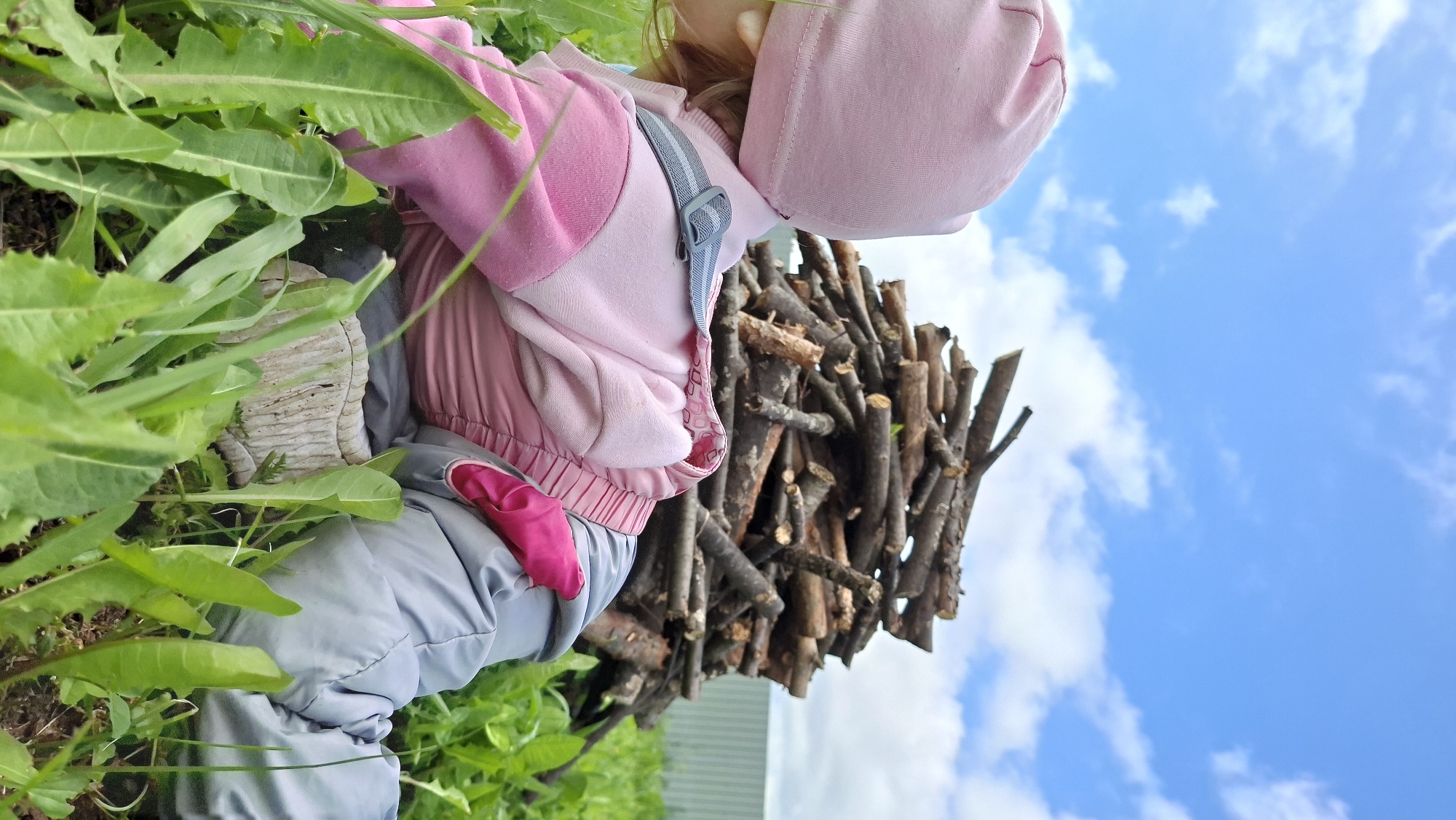 An outdoor image, taken from a low angle and rotated counter-clockwise, shows a child engaging with a natural environment. The child, clad in a pink hooded jacket or hoodie, a pink hat, and grey pants, is seen from a partial back and side view, bending down or crouching amidst vibrant green foliage and grass in the foreground. A grey strap, possibly from a backpack or carrier, is visible over their shoulder. To the right of the child, a substantial, tall pile of neatly cut wooden branches or logs stands vertically. The upper portion of the image reveals a clear blue sky dotted with fluffy white clouds, indicating a bright, sunny day. The scene suggests a peaceful moment of outdoor play or exploration in a rural setting, consistent with the location of Большой Букор, Russia. No text is visible in the image.