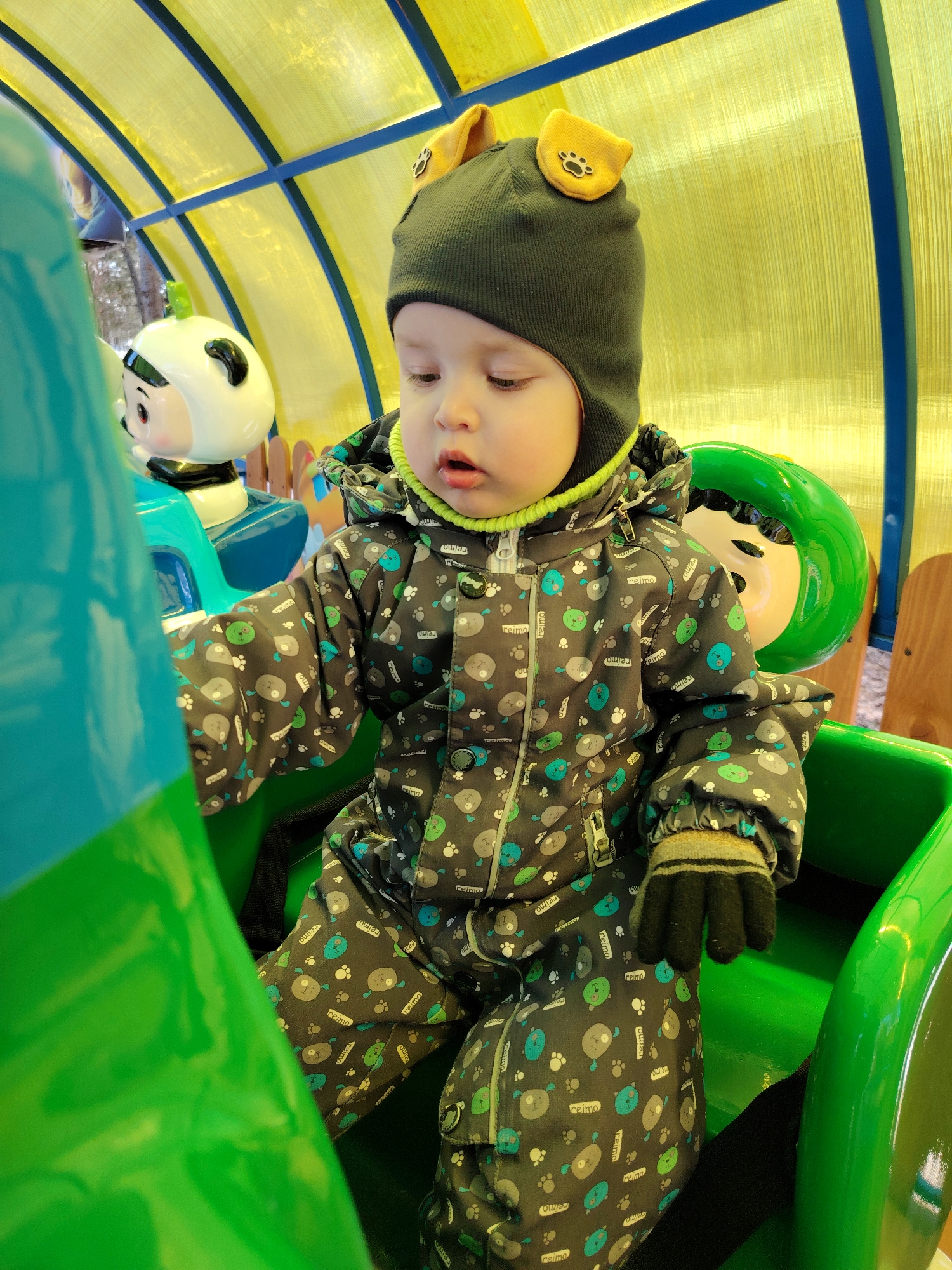 A young child in a patterned jumpsuit and a hat with animal ears is sitting in a brightly colored green seat, possibly part of a ride or play structure. The child is wearing a glove on one hand. The structure has a yellow translucent roof with blue supports. To the child's left is a blue and white cartoon character, and to the right is a green and white one. The location is Chaikovsky, Russia.