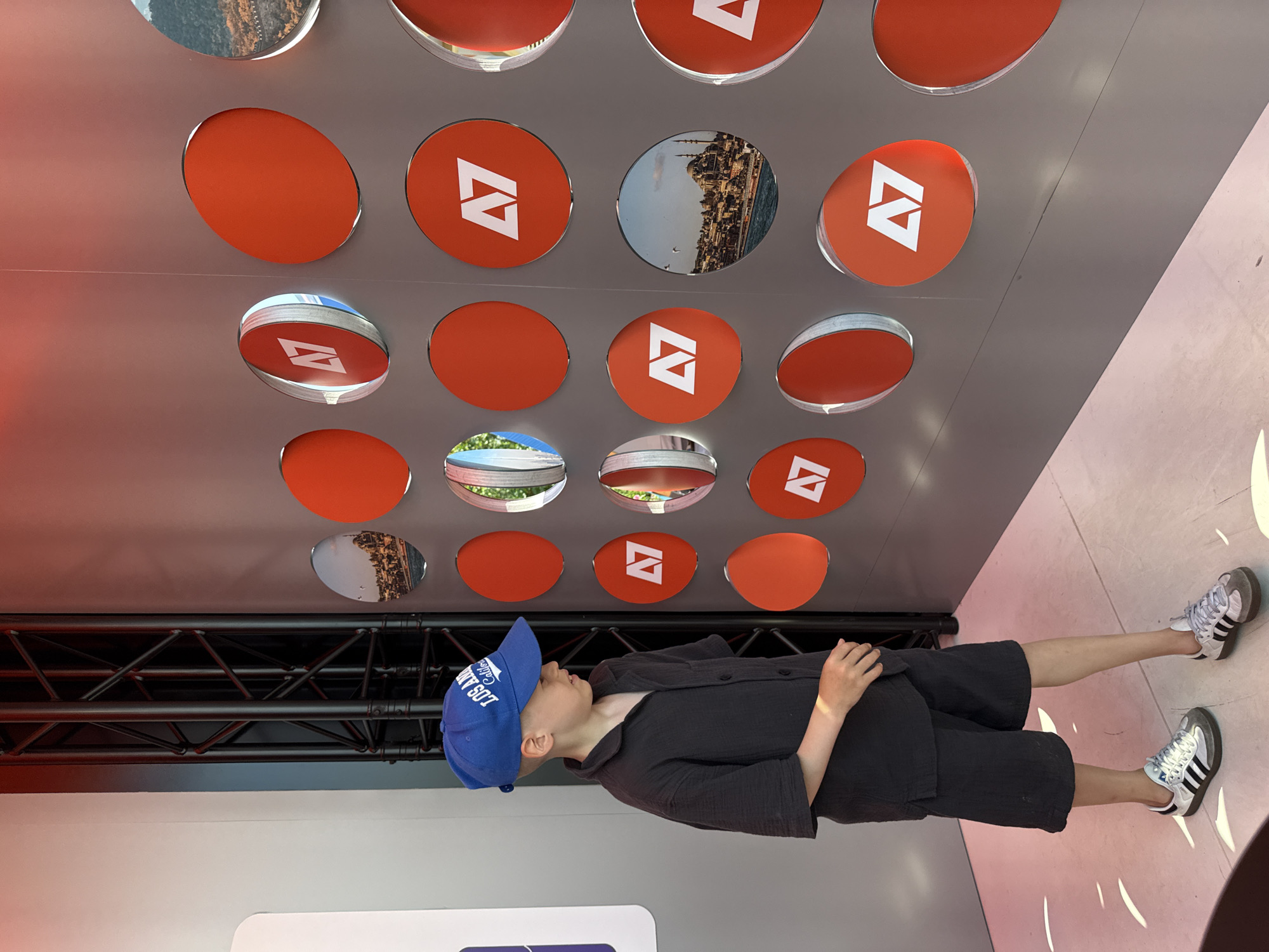 A young boy wearing a blue baseball cap and a dark grey short-sleeved shirt and shorts stands in front of a wall with circular cutouts. The cutouts are filled with either solid orange circles or circular images. Some of the images show cityscapes with mosques and bodies of water. The boy is looking to his right, towards the wall. He is wearing white sneakers with black stripes. Behind the boy, a black metal truss structure is visible. The floor is tiled and reflects the lighting. The overall scene appears to be an exhibition or a public space. The location is identified as Moscow, Russia.