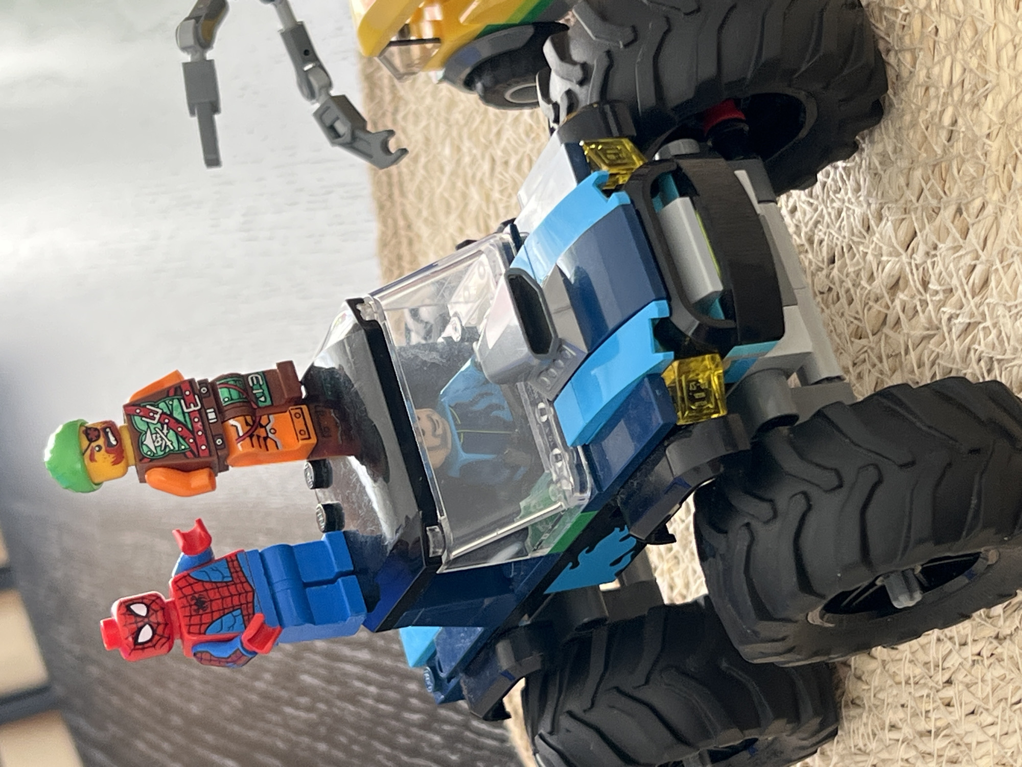 The image shows a close-up of Lego toys, specifically a monster truck and two minifigures, a zombie and Spider-Man. The monster truck is blue and black with large, textured tires. It has a transparent cockpit, through which a minifigure wearing a blue suit can be seen. Gold Lego elements are visible on the truck's exterior.
One minifigure, dressed as a zombie with green hair and a distressed brown and orange outfit, is positioned on the left side of the truck. Below it, Spider-Man, in his iconic red and blue suit, is also depicted as a minifigure. The toys are arranged on a textured, woven surface, possibly a rug or mat, with a light-colored background that suggests an indoor setting. A partial view of Lego robotic arms is visible in the upper left corner of the image. The lighting is bright, indicating daytime. There is no visible text in the image.