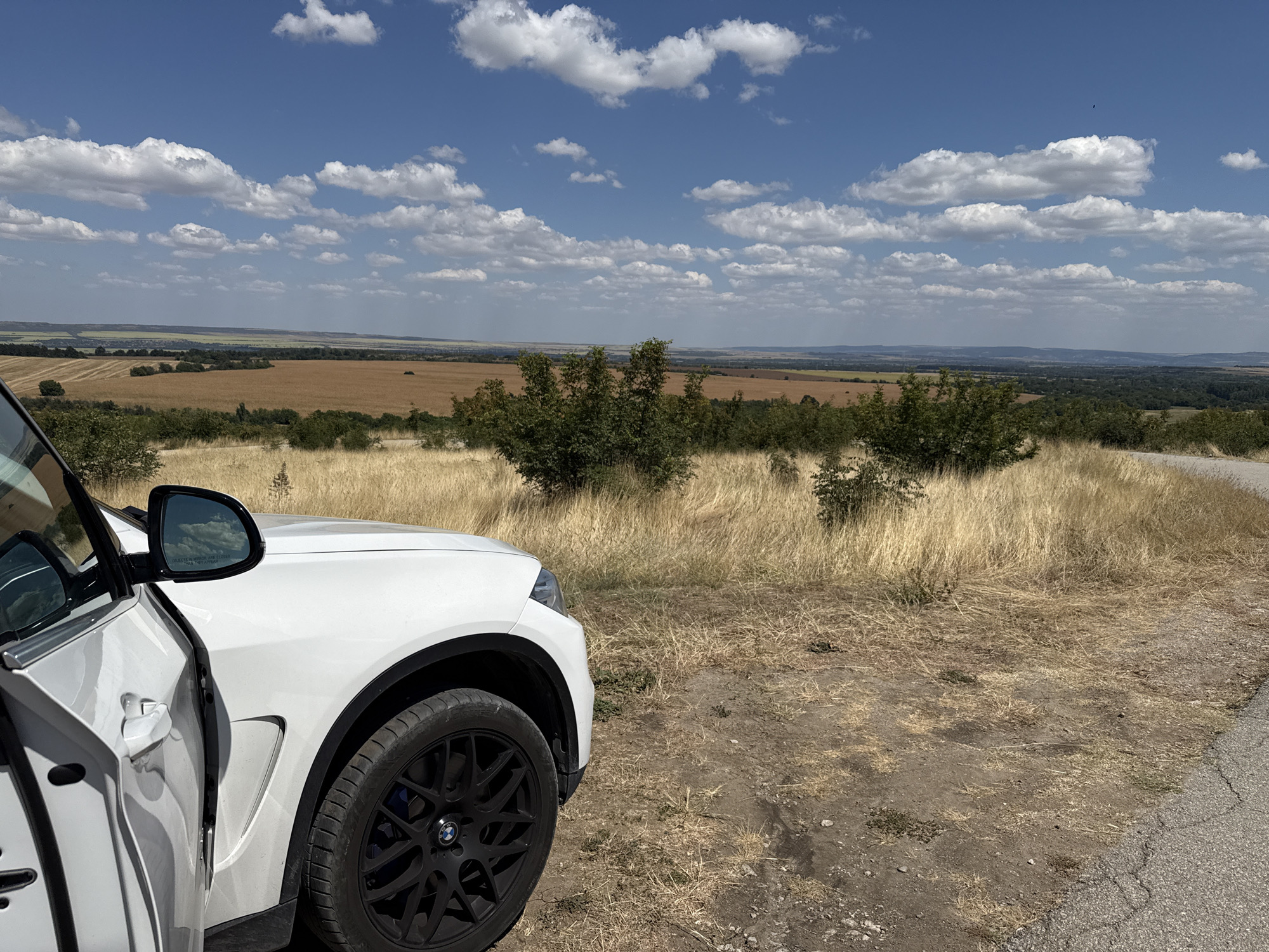 The image shows a white car parked on a dirt road overlooking a valley in Hotnitsa, Bulgaria. The car is partially visible on the left side of the frame, with its driver's side door open. The vehicle is a modern SUV with black rims and a blue brake caliper visible.
The landscape beyond the car is a rolling valley with agricultural fields, appearing mostly brown and golden, indicative of a dry season or harvested crops. Some scattered trees and patches of greenery are visible throughout the valley. In the background, the horizon is lined with distant hills and a hazy sky.
The sky is blue with scattered white, fluffy clouds, suggesting a clear, sunny day. The foreground is dominated by dry, tall grass and sparse vegetation, characteristic of a rural or steppe-like environment. A portion of an asphalt road is visible on the right side of the frame. The scene is static and appears to be a scenic viewpoint.