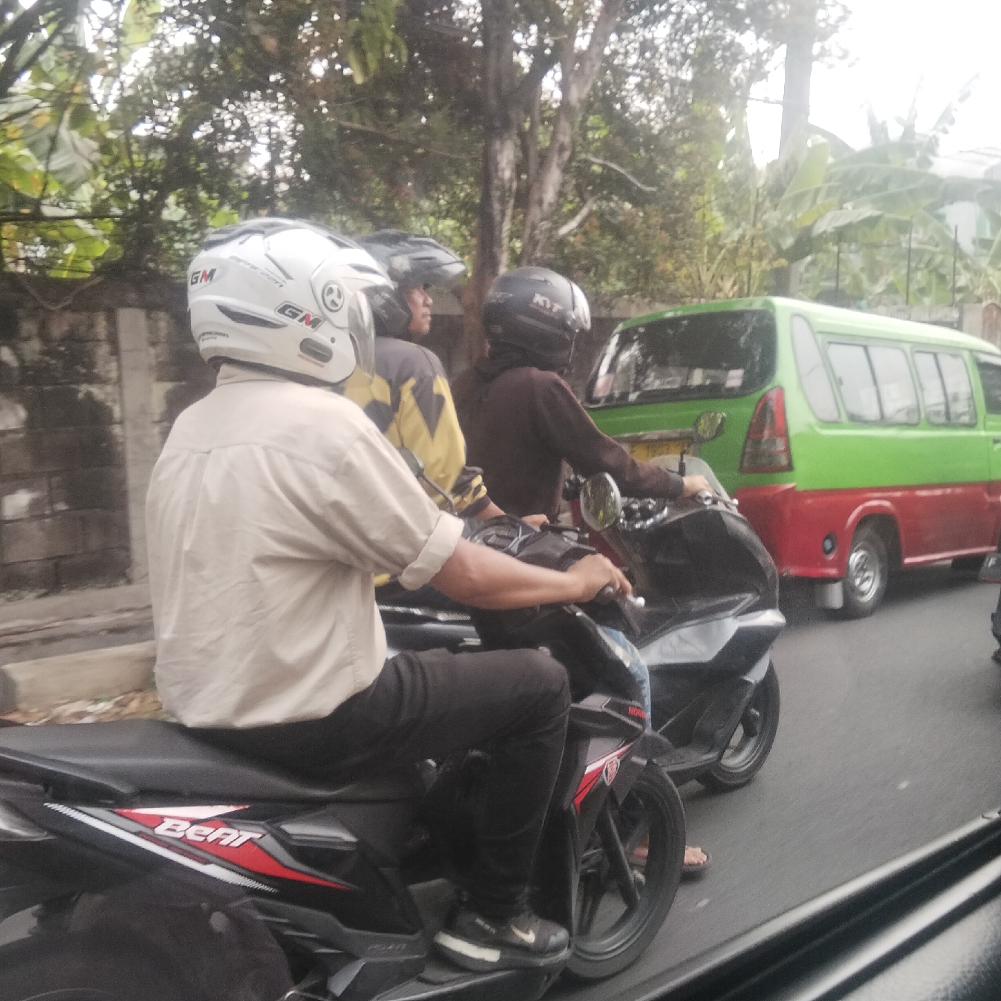 The image shows a street scene in Bogor, Indonesia. Two people are riding motorcycles. The person in the foreground is wearing a light-colored shirt and black pants, with a white helmet. They are on a black motorcycle with a red and white "BEAT" logo. The person behind them is wearing a dark jacket and a black helmet, also on a motorcycle. In the background, a green and red van is visible. The environment appears to be a moderately busy street with trees and buildings.
