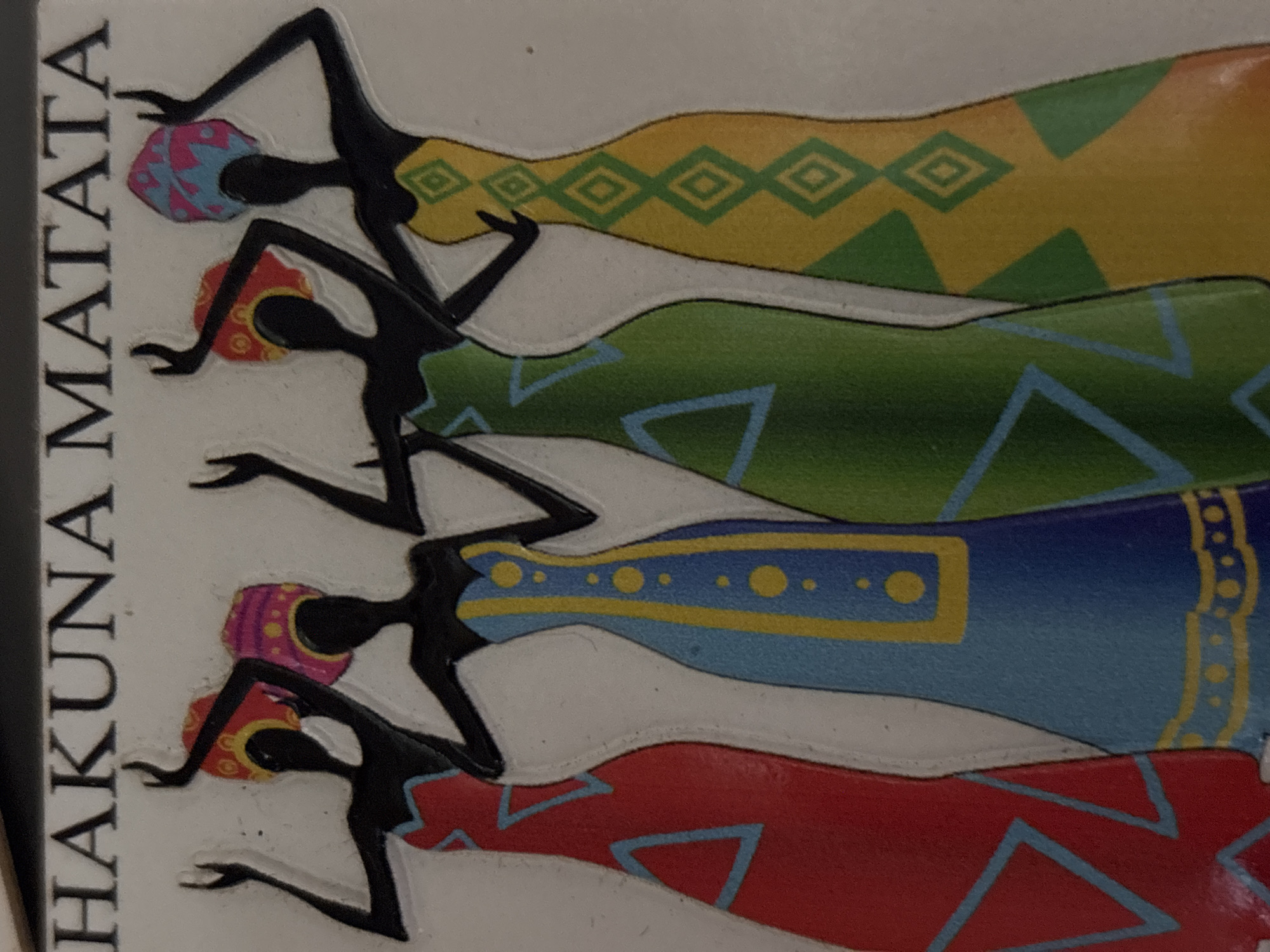 The image depicts a decorative object or piece of art featuring four stylized silhouettes of women in colorful dresses. The women are arranged in a vertical stack, each wearing a different patterned dress in colors such as yellow, green, blue, and red. The figures are black silhouettes, and they are each wearing a patterned head covering. To the left of the figures is the text "HAKUNA MATATA" written vertically. The background is white.